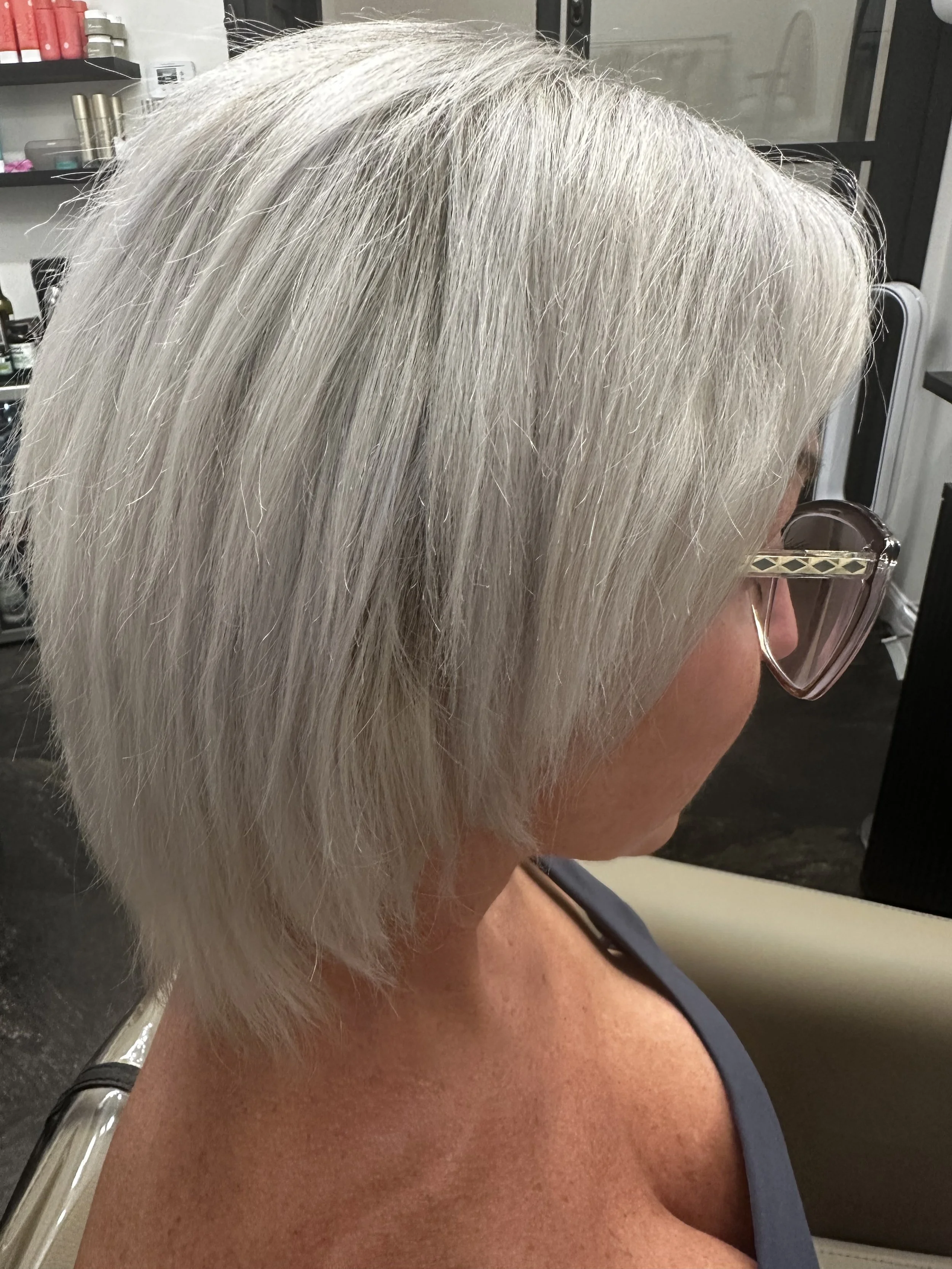 Close-up of a woman with short, silver-gray hair wearing glasses, sitting in a salon or similar setting.