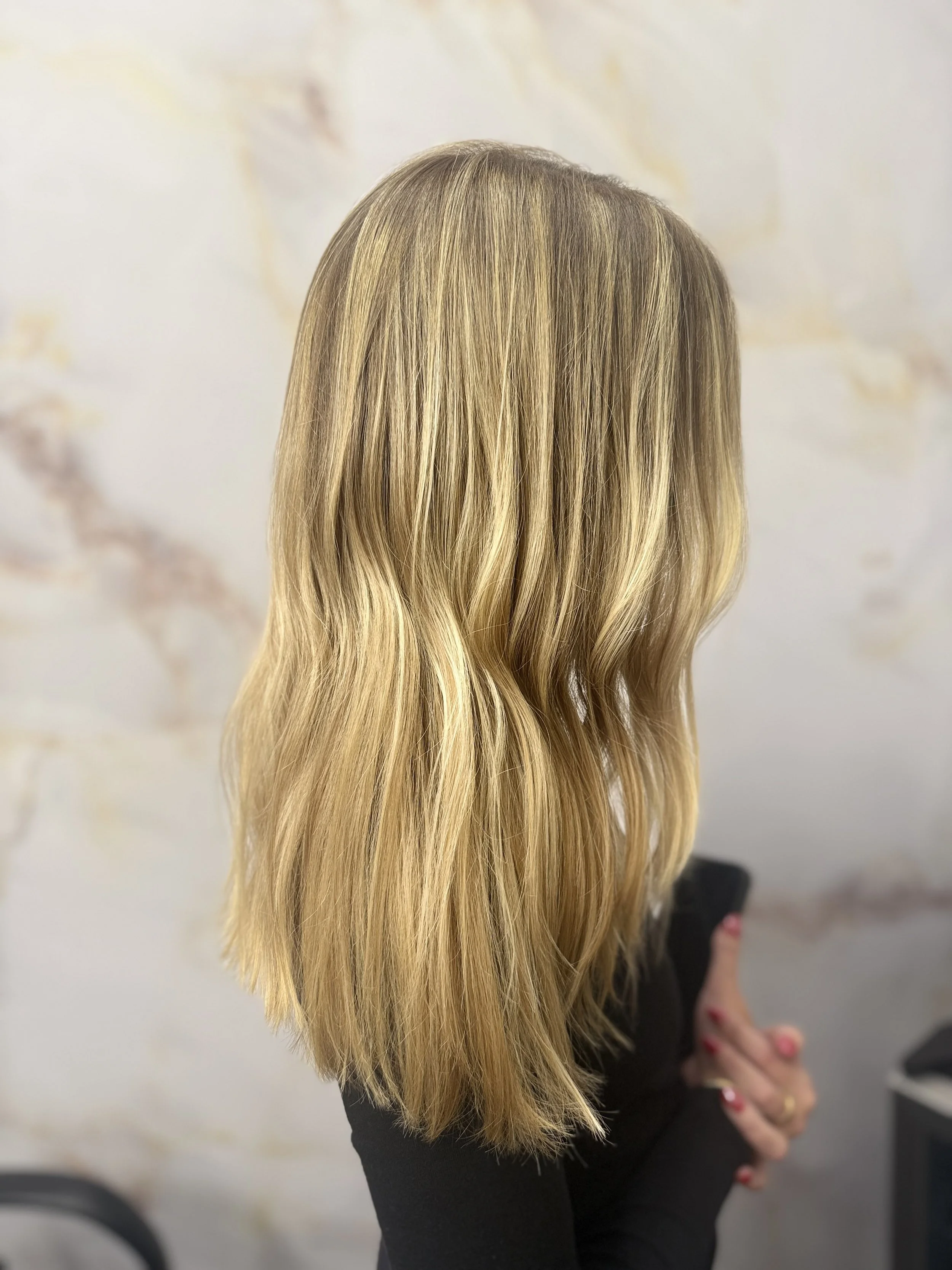 Back view of a woman with long, wavy blonde hair, wearing a black top, standing in an indoor space with a light-colored wall.
