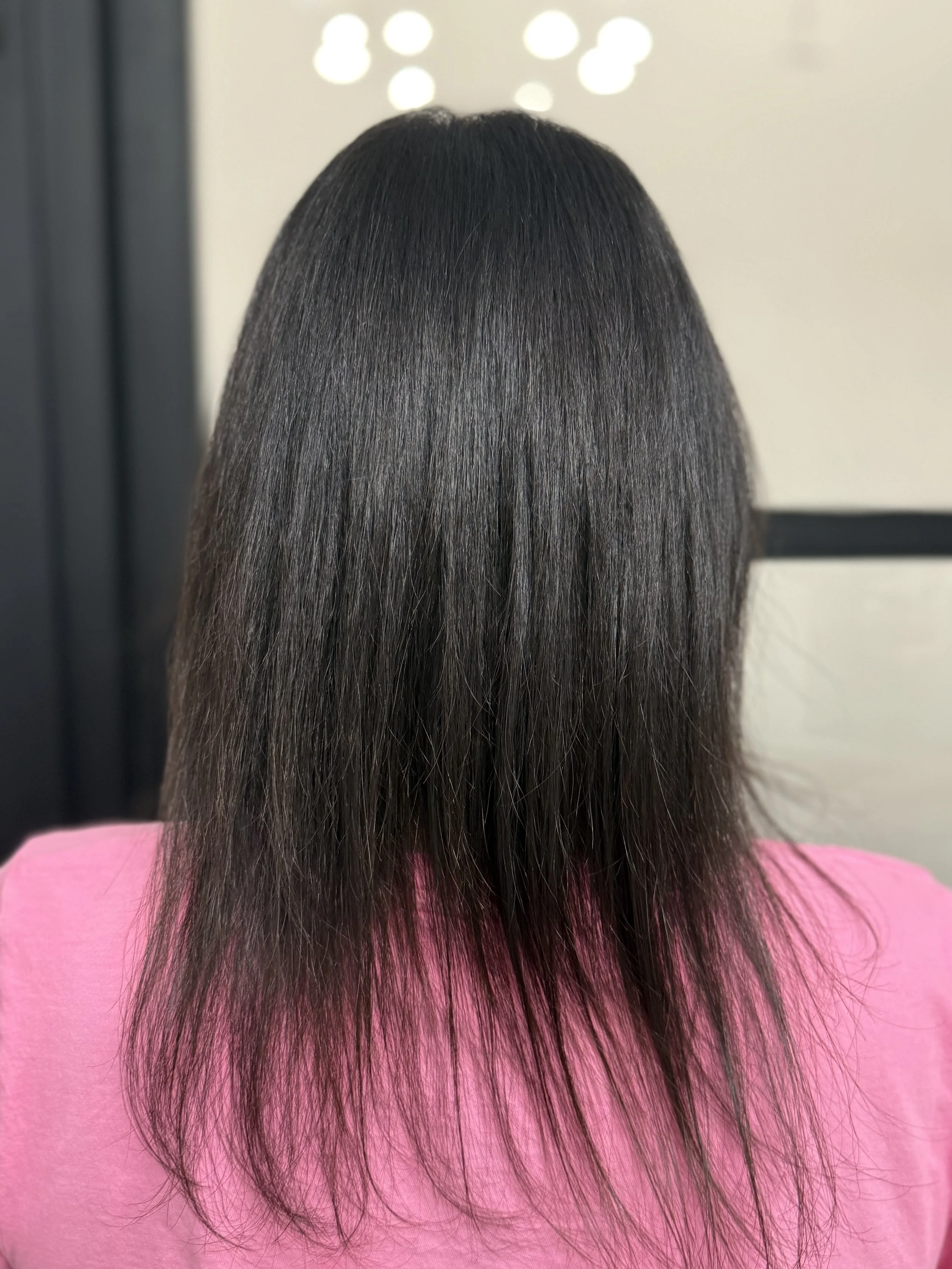 The image shows the back of a person with dark, straight, and shiny hair that is longer in the back, wearing a pink top.
