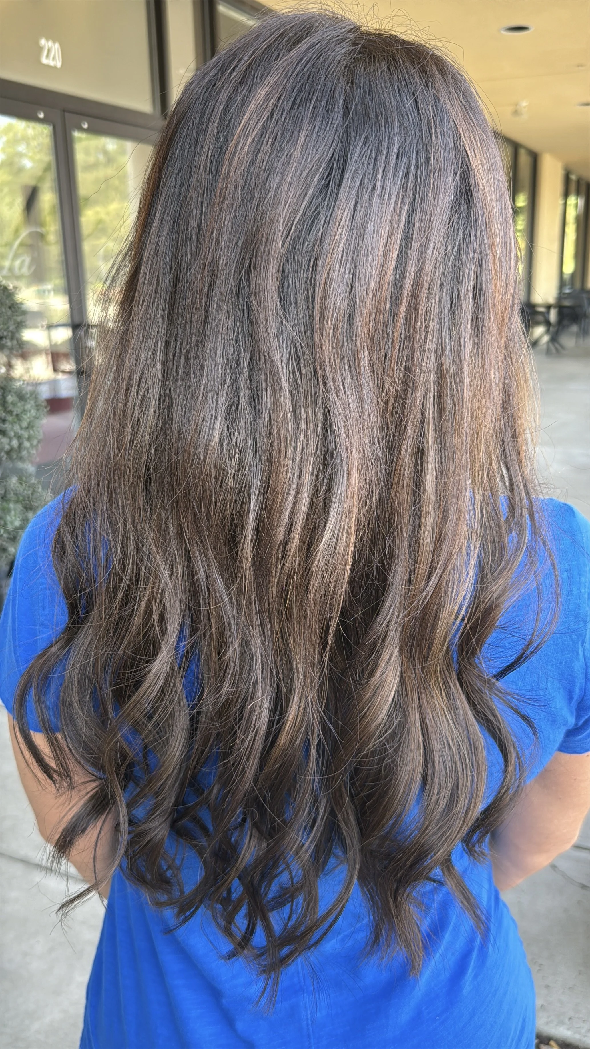 Back of woman with long wavy hair wearing a blue shirt standing outside near a glass window
