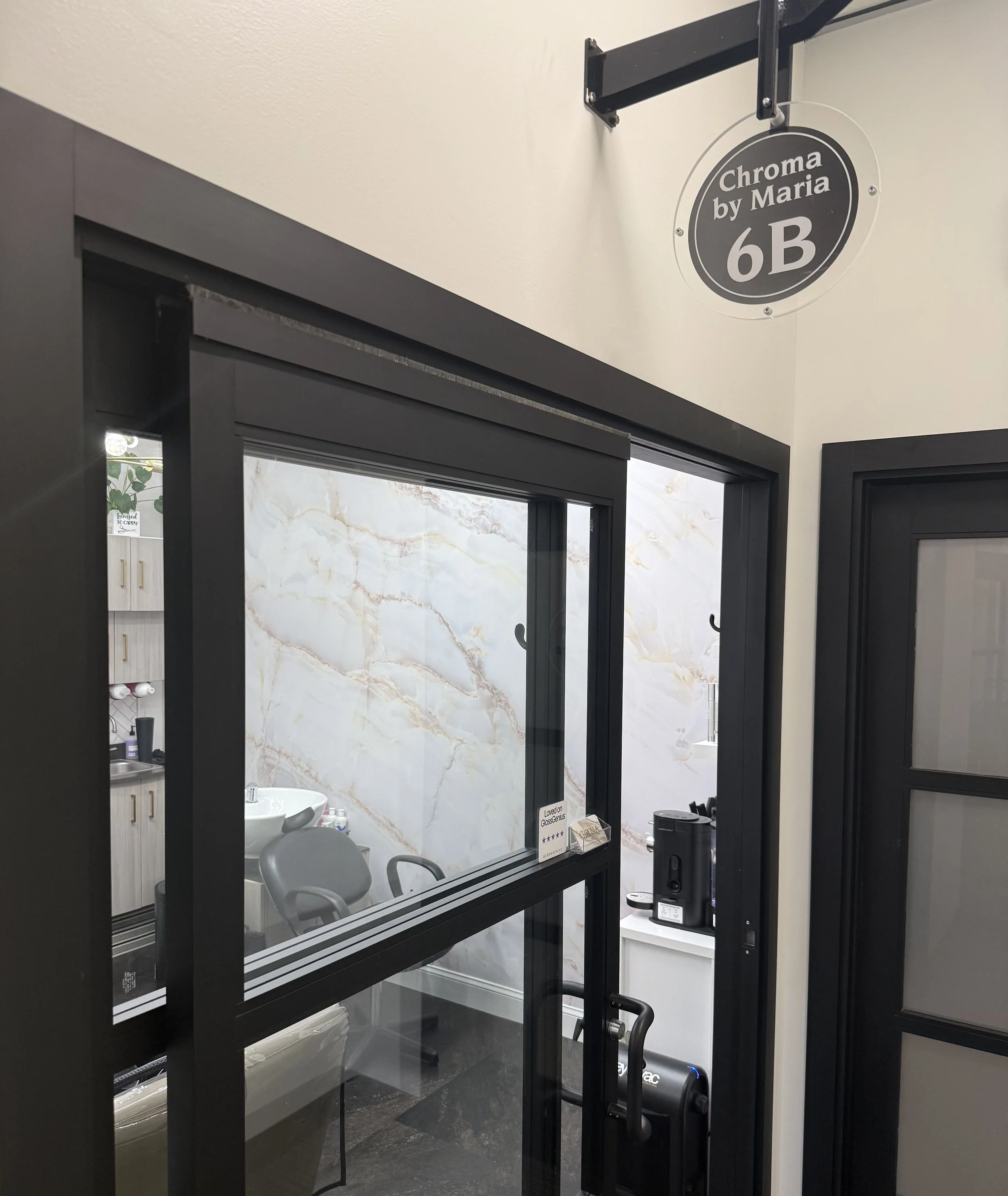 Interior of a modern building with a glass door, a white marble wall, a mirror, a sink, and a stool visible through the door. There is a sign indicating a room or section labeled 'Chroma by Maria 6B' hanging near the ceiling.
