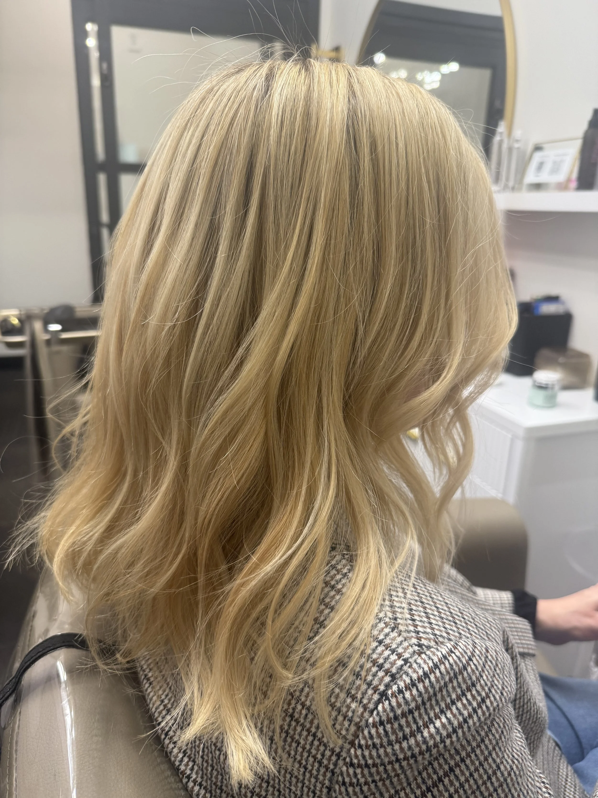 Blonde hair styled in loose waves, seen from behind, resting on the shoulders of a person wearing a houndstooth patterned blazer.