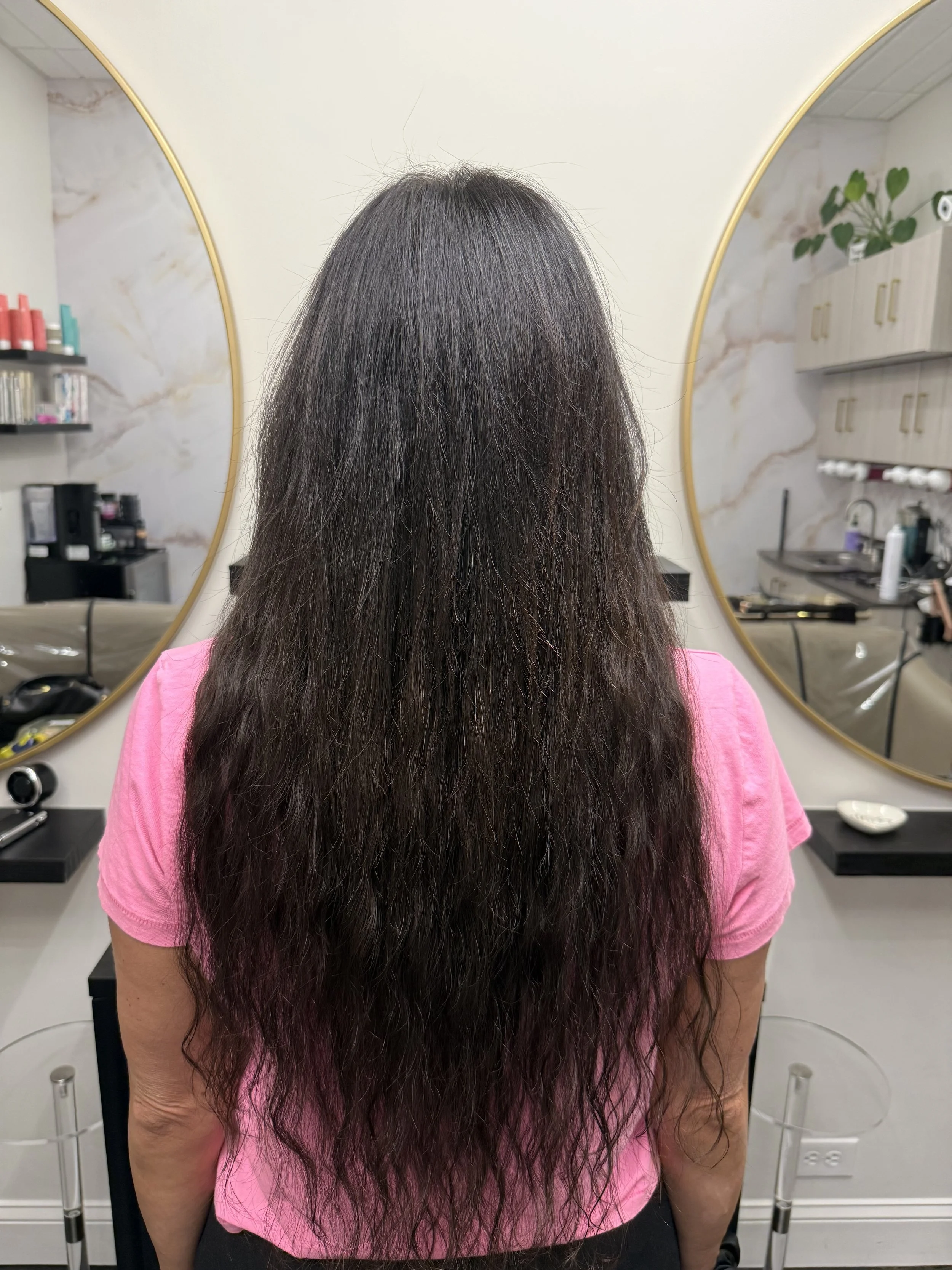 Back of a woman with long, wavy dark hair wearing a pink shirt, standing in front of a mirror in a salon. The salon has shelves with hair products and a marble wall background.