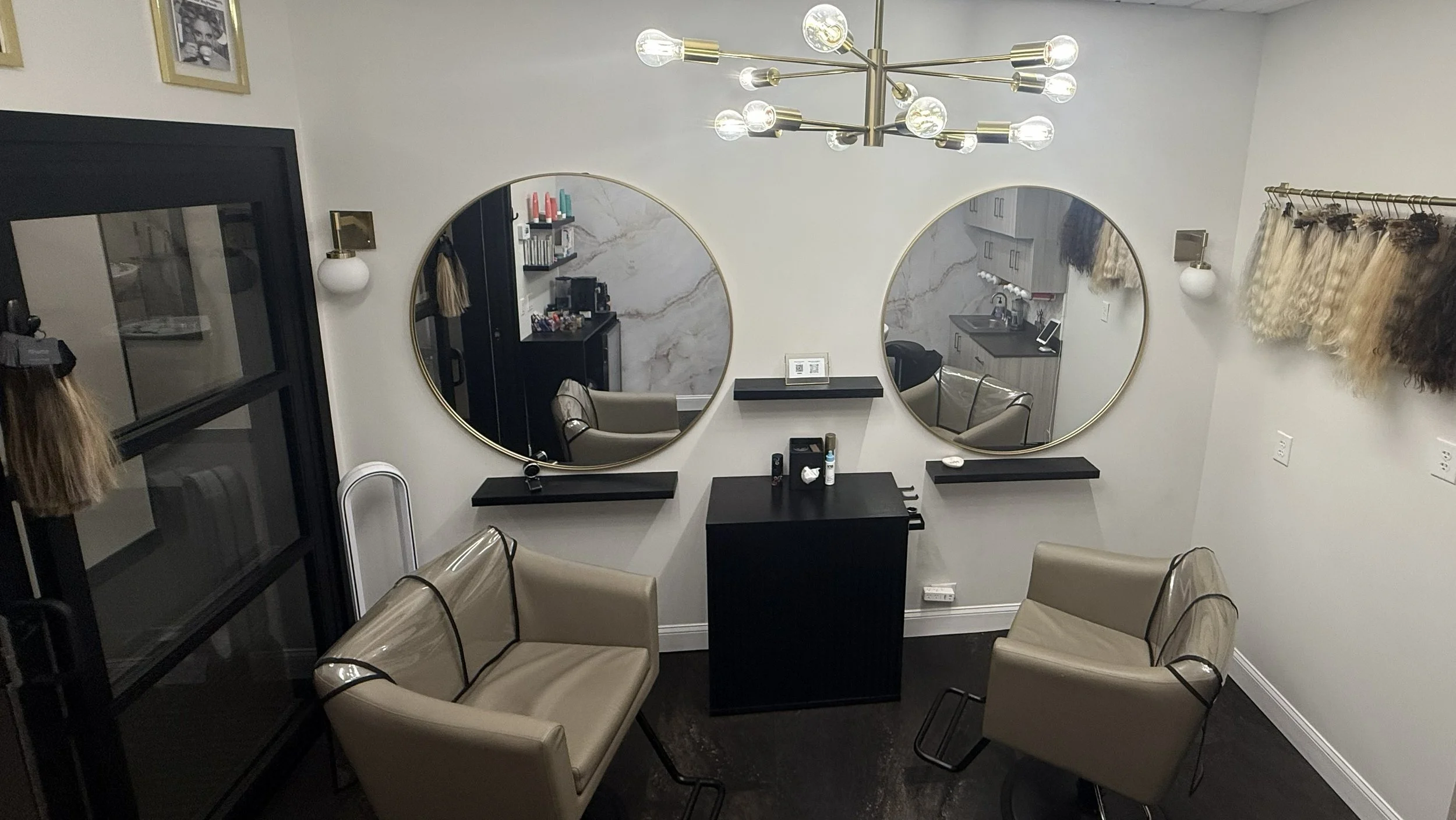 Salon with beige chairs, a black desk with supplies, and two large round mirrors on the wall, decorated with a modern chandelier and a wall of hair extensions on the Philadelphia Mainline - Wayne Pa