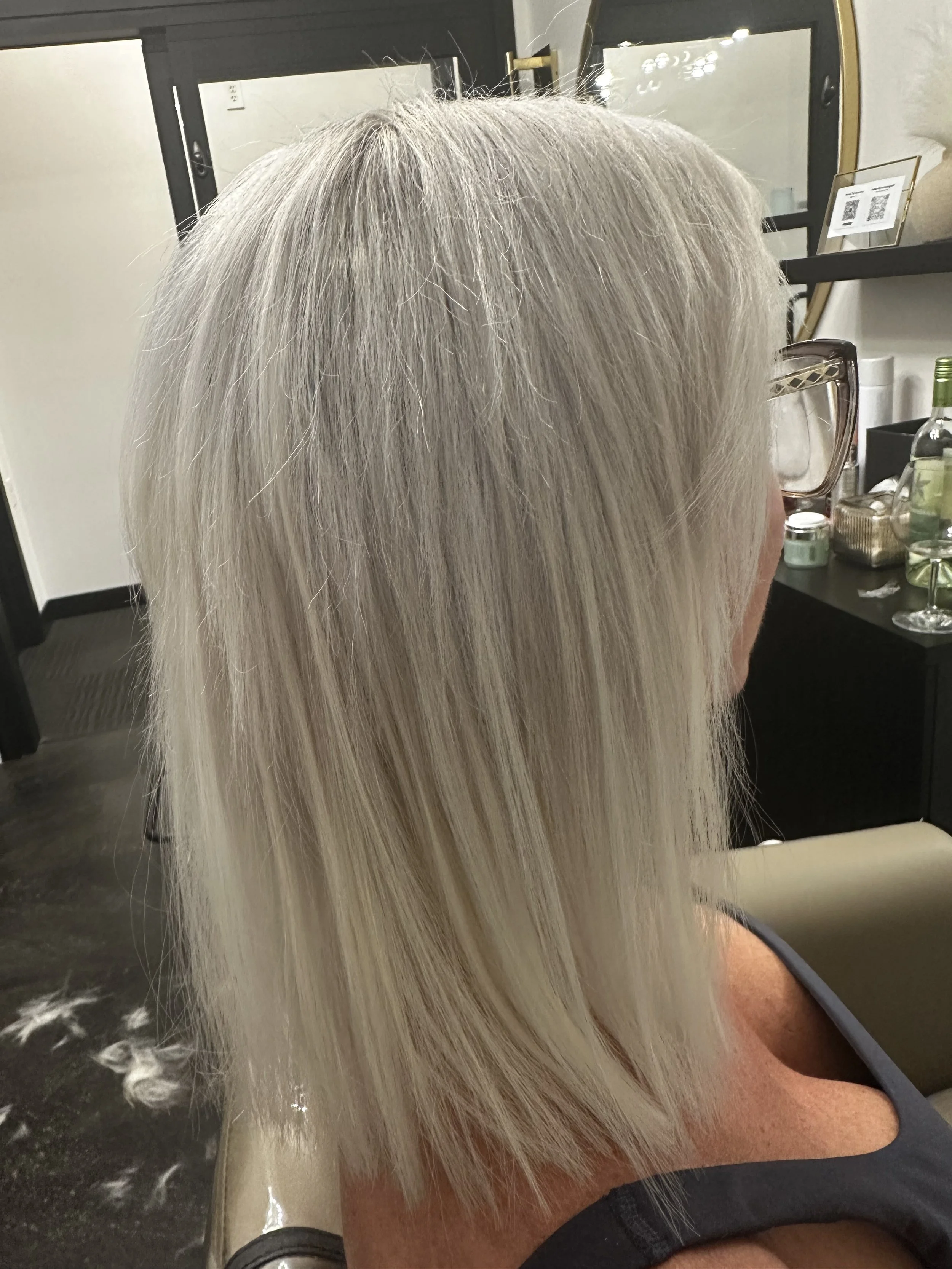 The image shows a person with long, straight, platinum blonde hair sitting at a vanity mirror in a salon or similar setting, with various salon tools and products on the counter.