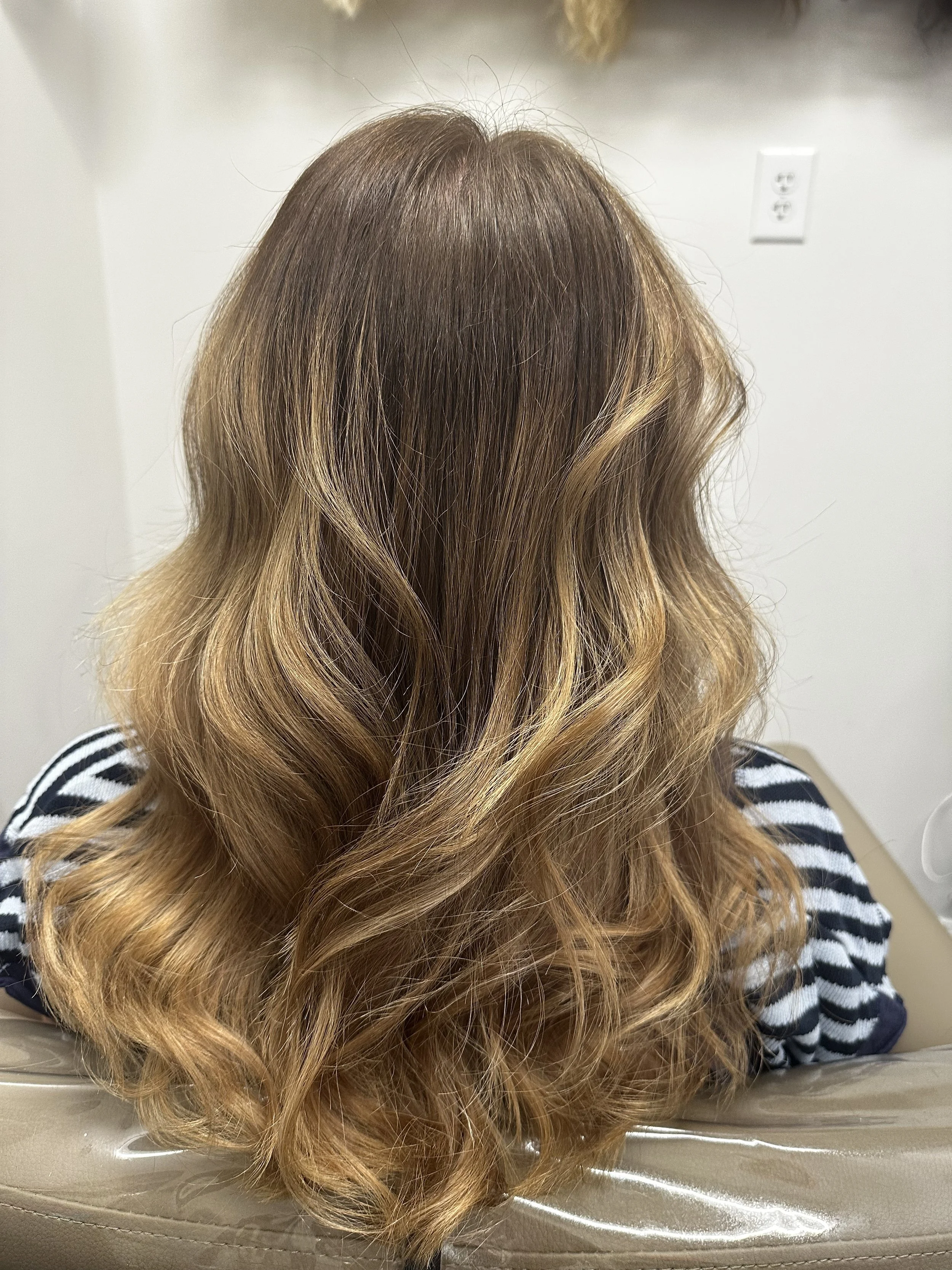 The back of a person's head with long, wavy, light brown hair that has blonde highlights. The person is wearing a black and white striped shirt and is sitting on a beige chair or sofa.