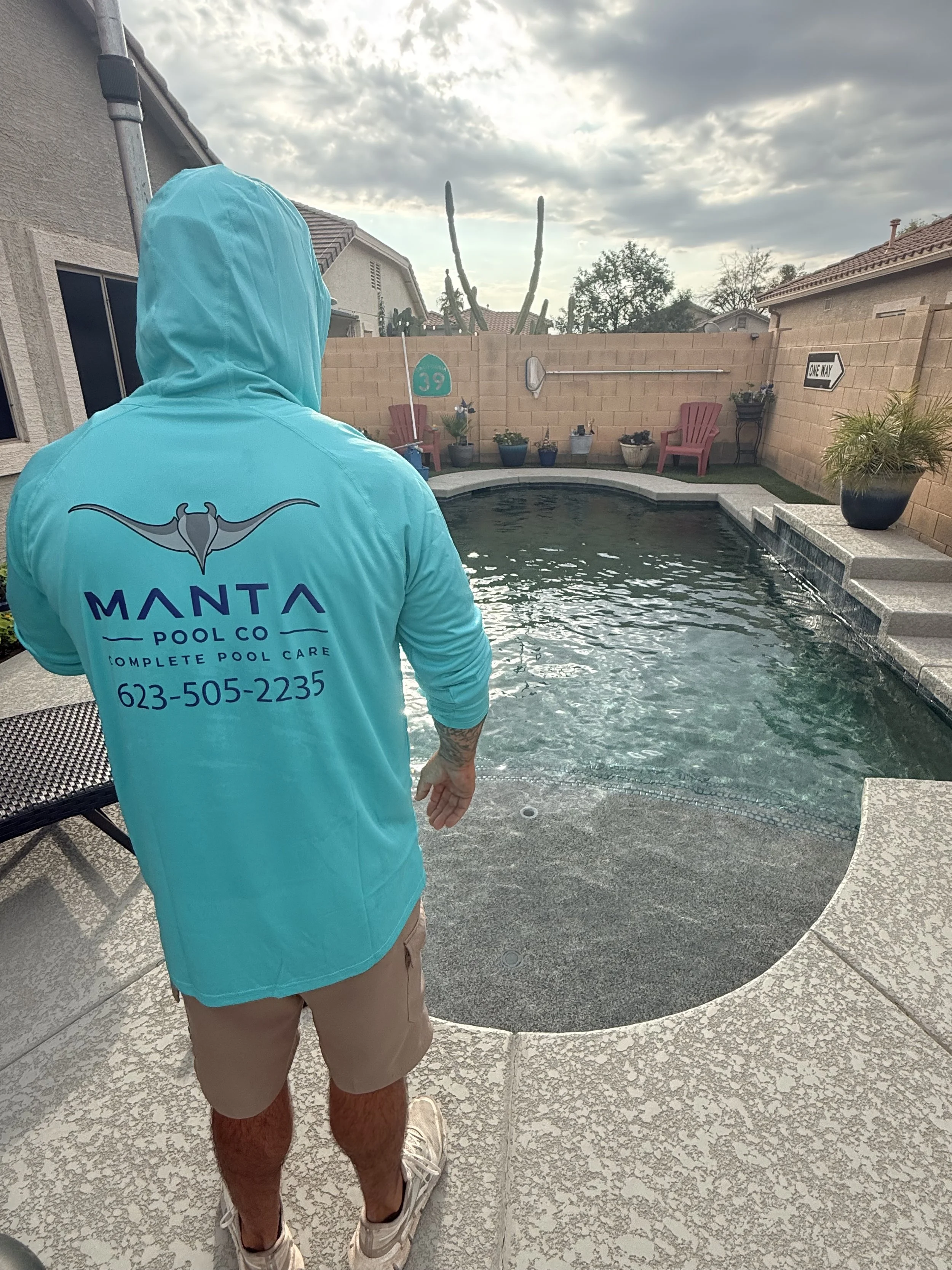 Person standing by a backyard pool, wearing a turquoise hoodie with a manta ray logo and text for a pool cleaning company, beige shorts, and sneakers, with a cloudy sky and cactus plants in the background.