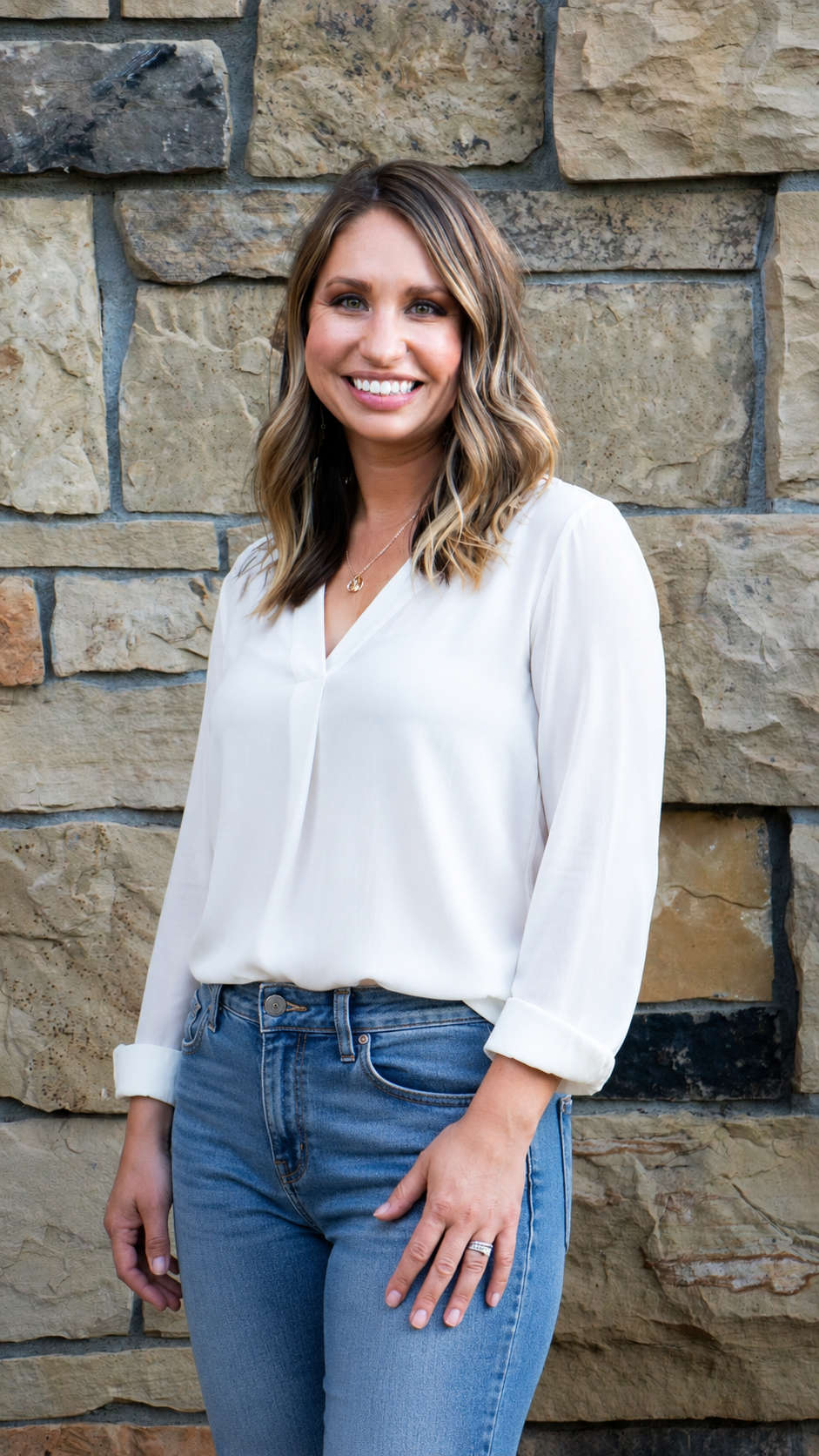 Owner of Petals + Bloom smiling, wearing a white blouse and blue jeans, standing in front of a stone wall.