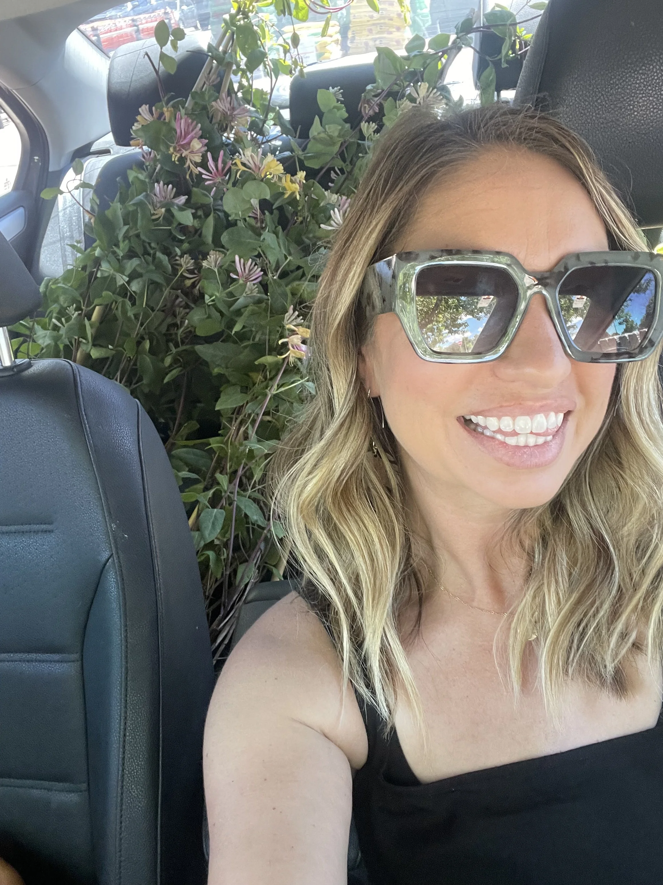 Owner of Petals + Bloom wearing large silver sunglasses smiling, sitting in the backseat of a car with a large bush with purple and yellow flowers behind her.