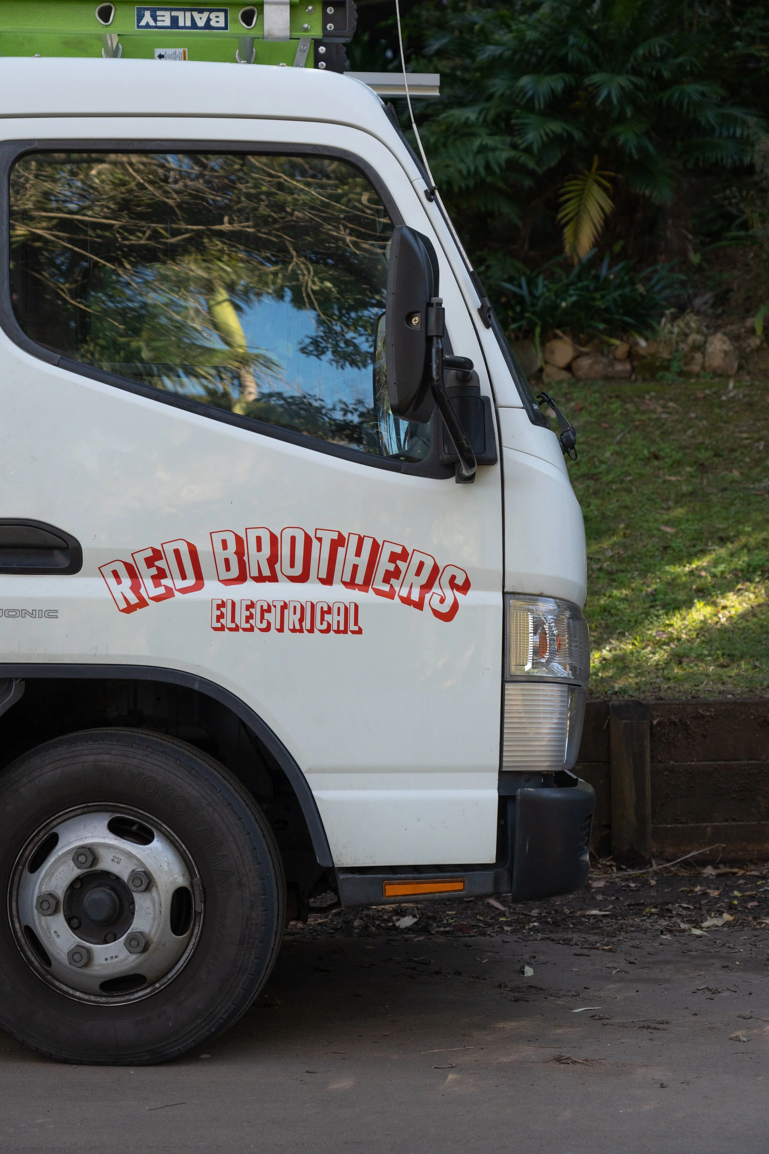 Close-up of a white utility van with a company logo 'Red Brothers Electrical' on the side, parked on a street with a background of greenery. The van has a large black side mirror, a visible wheel, and part of a green ladder on top.