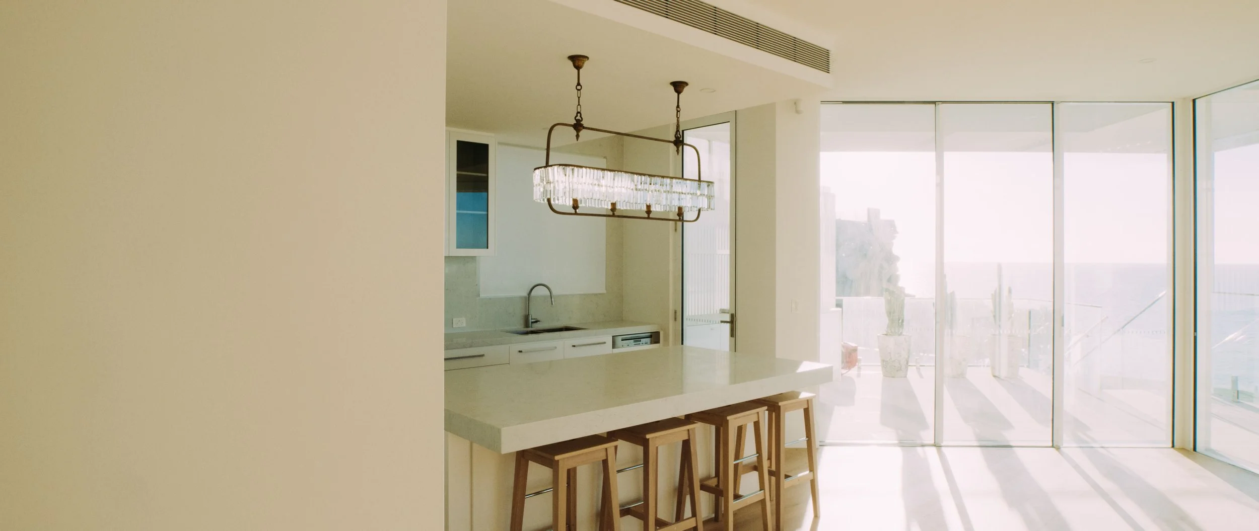 Modern kitchen with a white island, wooden bar stools, a chandelier, and large glass sliding doors leading to a balcony with potted plants and city view