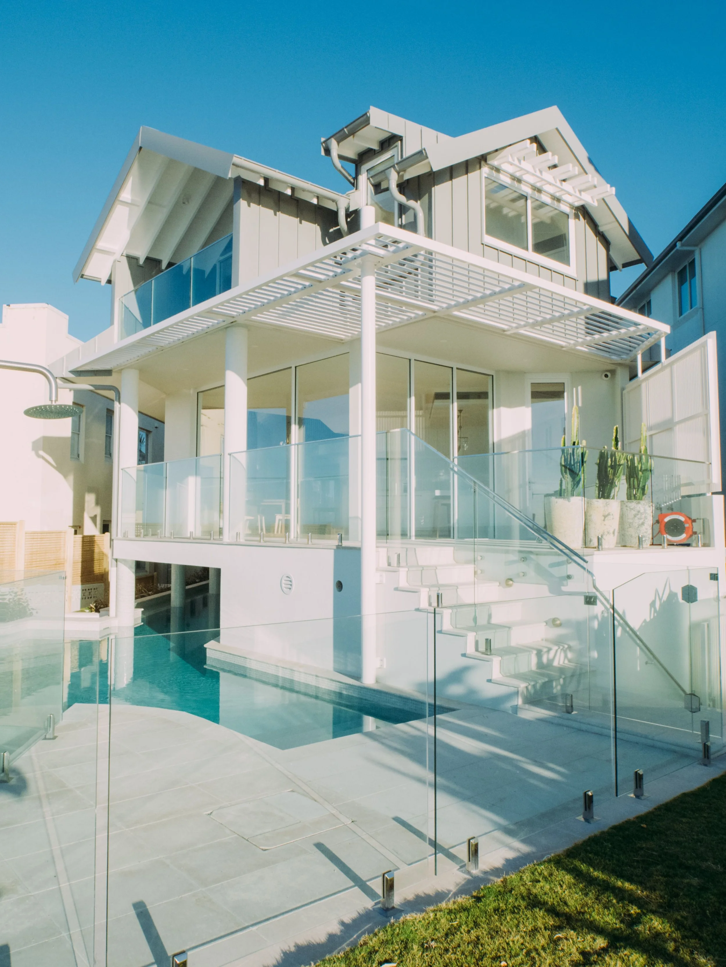 Modern house with glass balcony, a swimming pool, and outdoor shower, under a clear blue sky.