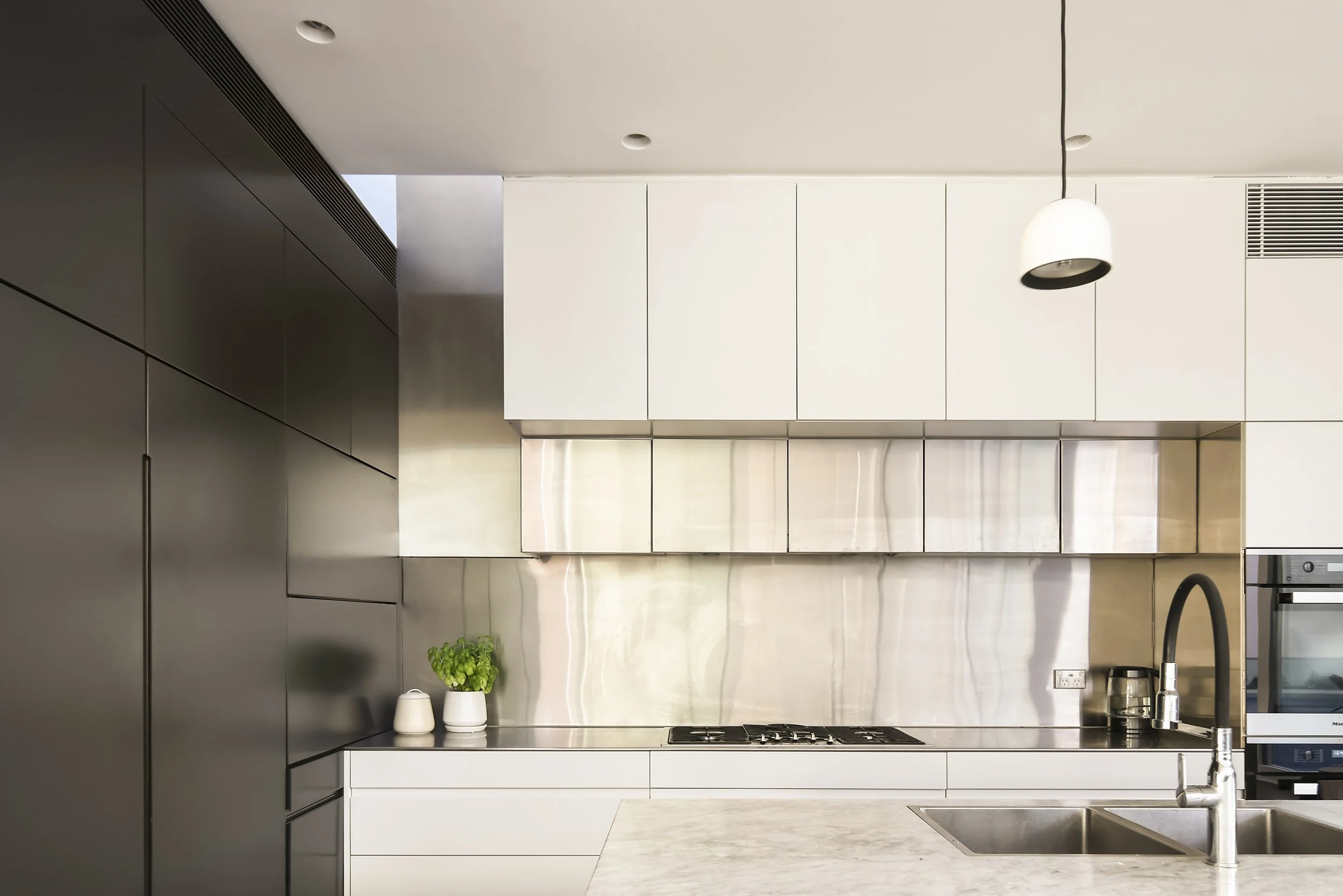 Modern kitchen with black and white cabinets, stainless steel backsplash, a countertop with a marble finish, a small plant, two white cups, a stove, a sink with a faucet, and a hanging white pendant light.