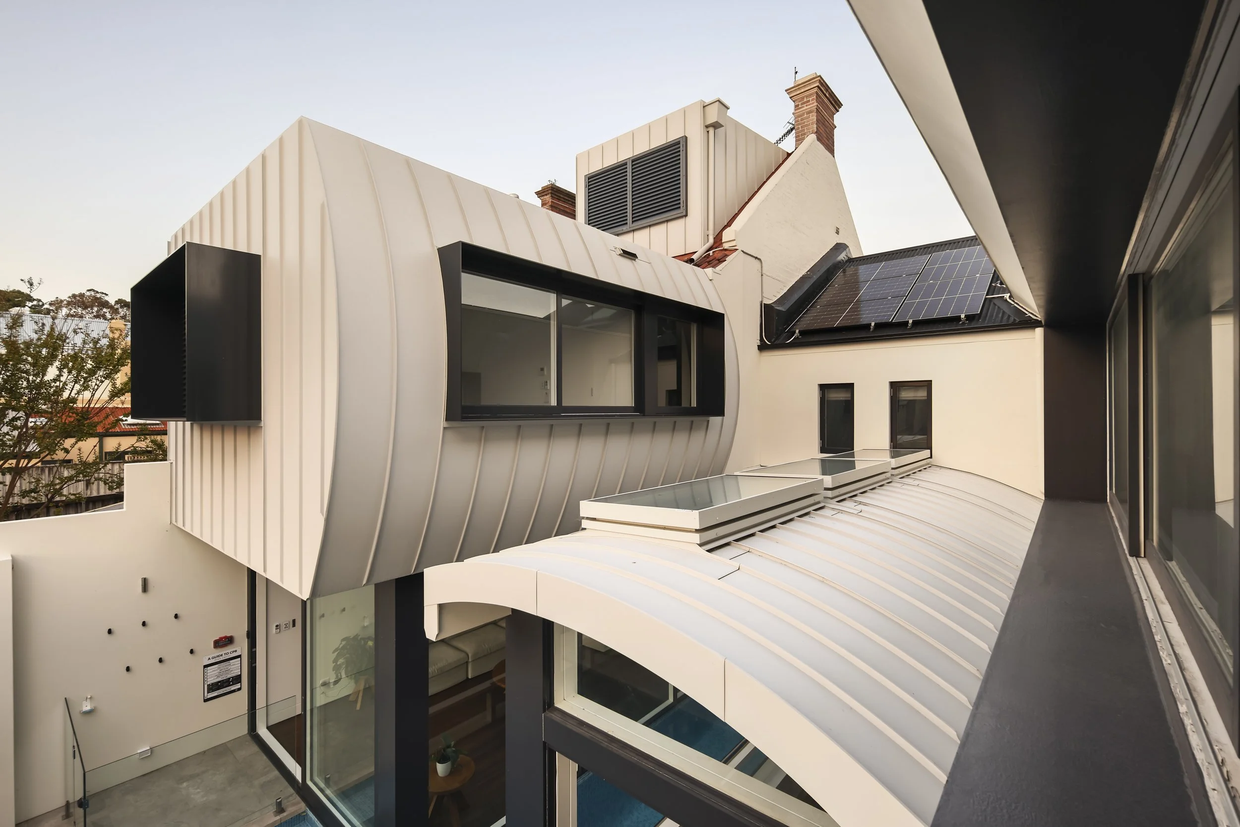 View of a modern residential building with a white, curved metal exterior, large windows, and solar panels on the roof, with neighboring rooftops and trees in the background.