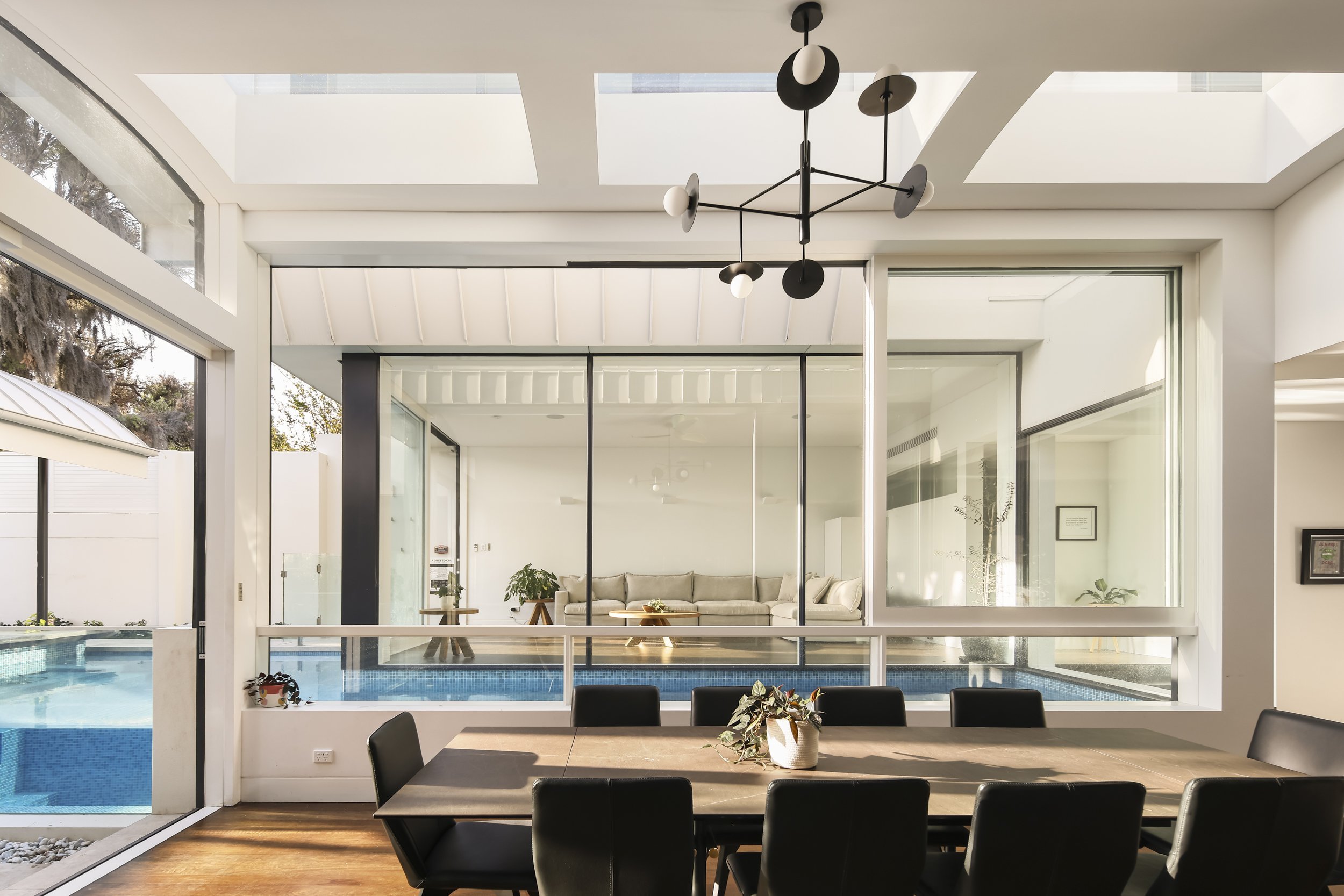 Modern dining room with black chairs around a rectangular wooden table, a white potted plant in the center, large glass windows and doors, view of a pool and outdoor area with trees, and a contemporary black and white chandelier hanging from the ceil