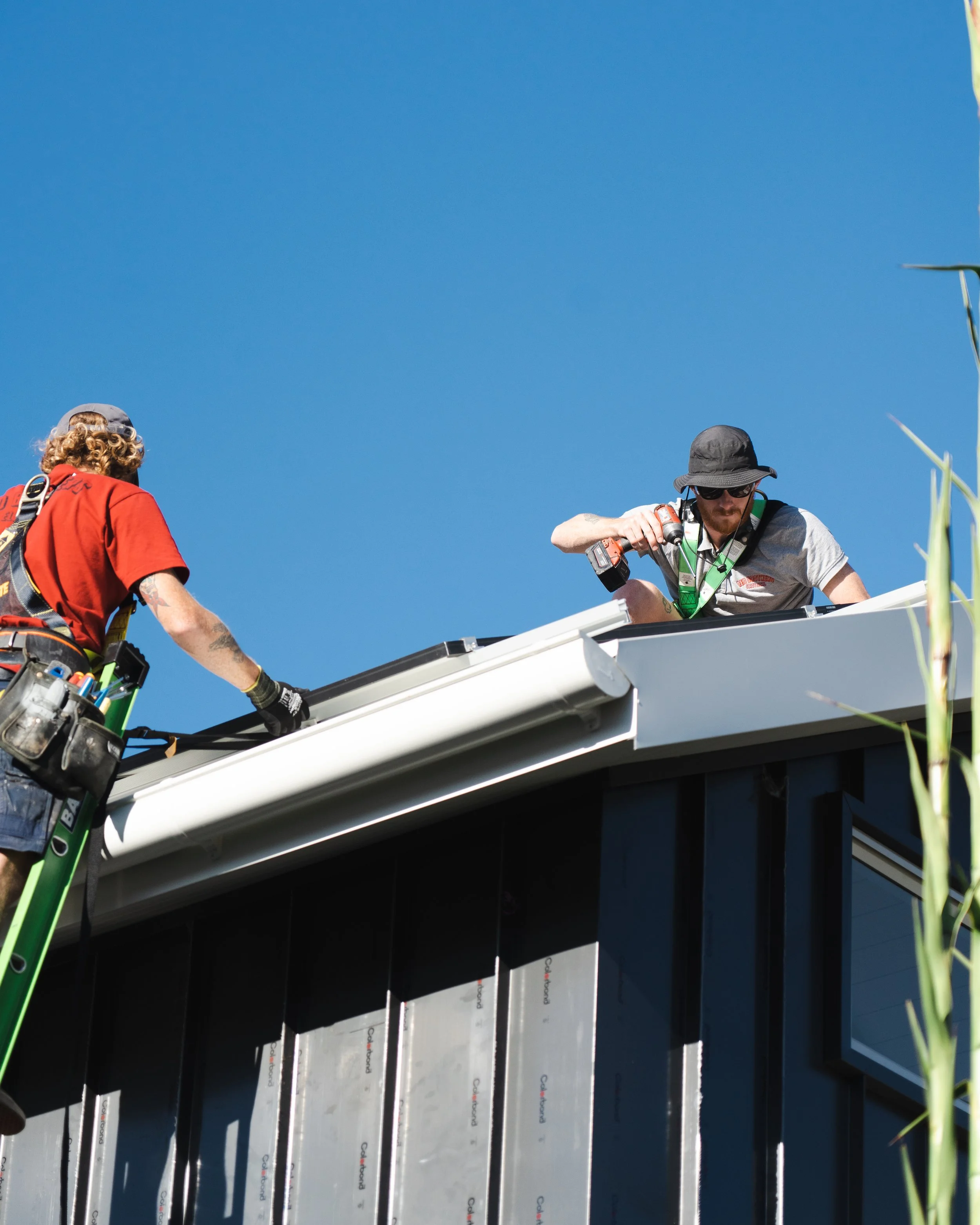 Two workers on a roof working with solar panel installation. One worker is using a cordless drill, and the other is assisting. The panels are mounted on the roof of a modern building, against a clear blue sky.