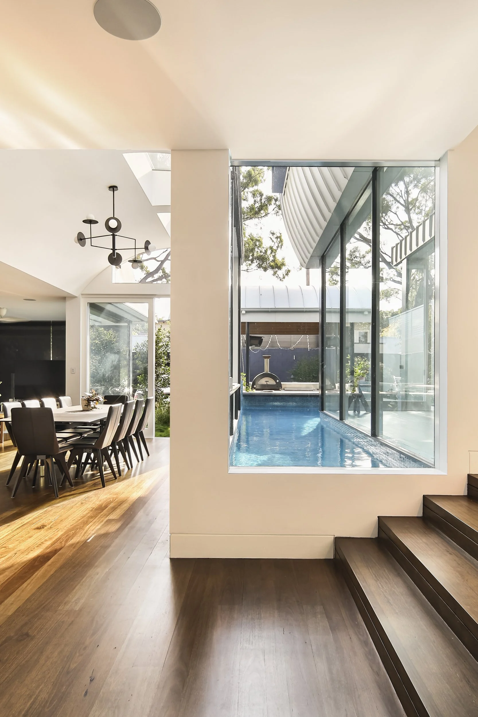Modern interior home with a dining area on the left, large windows and a sliding glass door leading to a pool area on the right, wooden floors, and contemporary light fixtures.