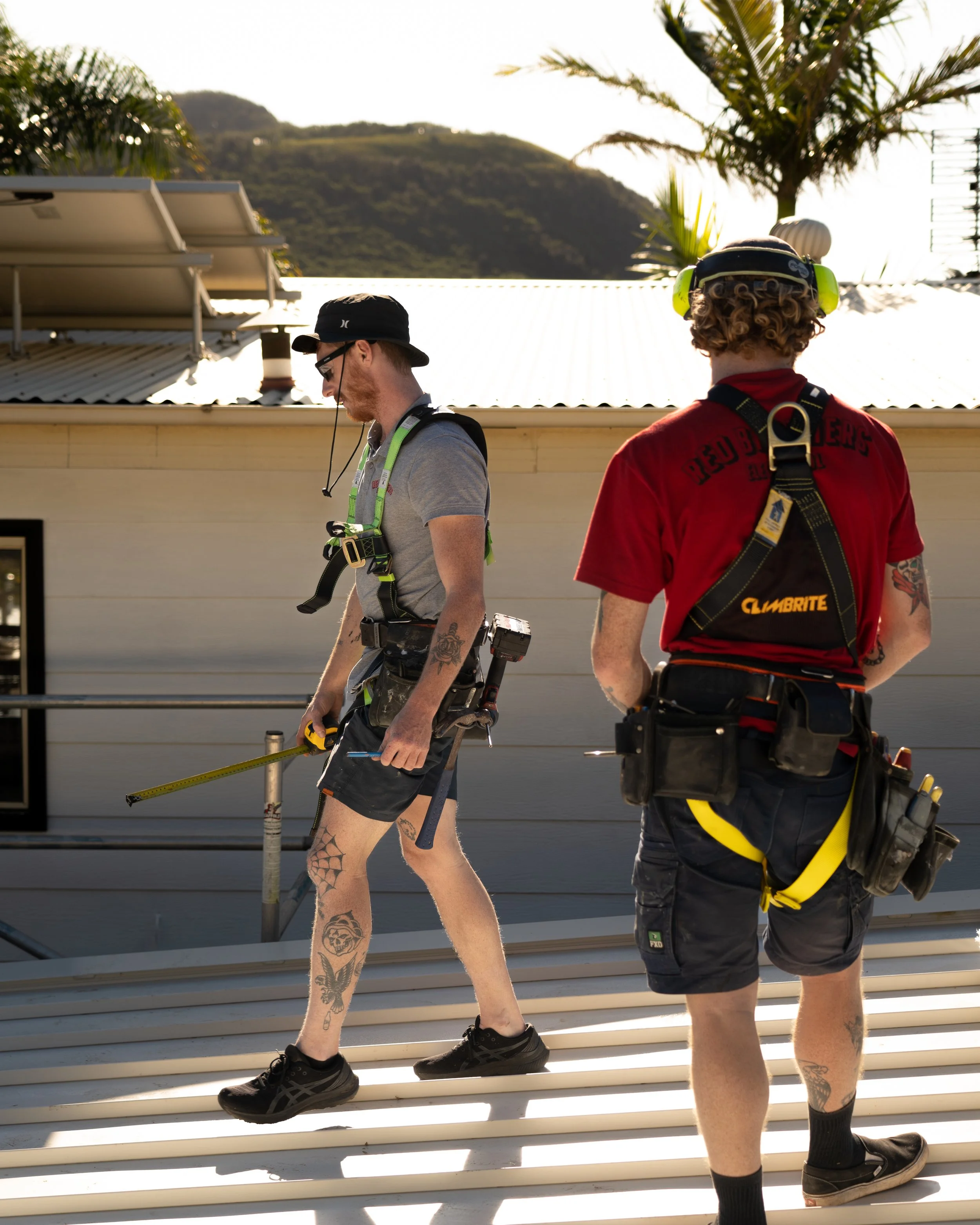 Two construction workers walking down angled metal stairs outdoors during daylight. The worker on the left wears a black hat, glasses, a gray t-shirt, and sports tattoos on his left leg. The worker on the right has curly hair, a black helmet with bri