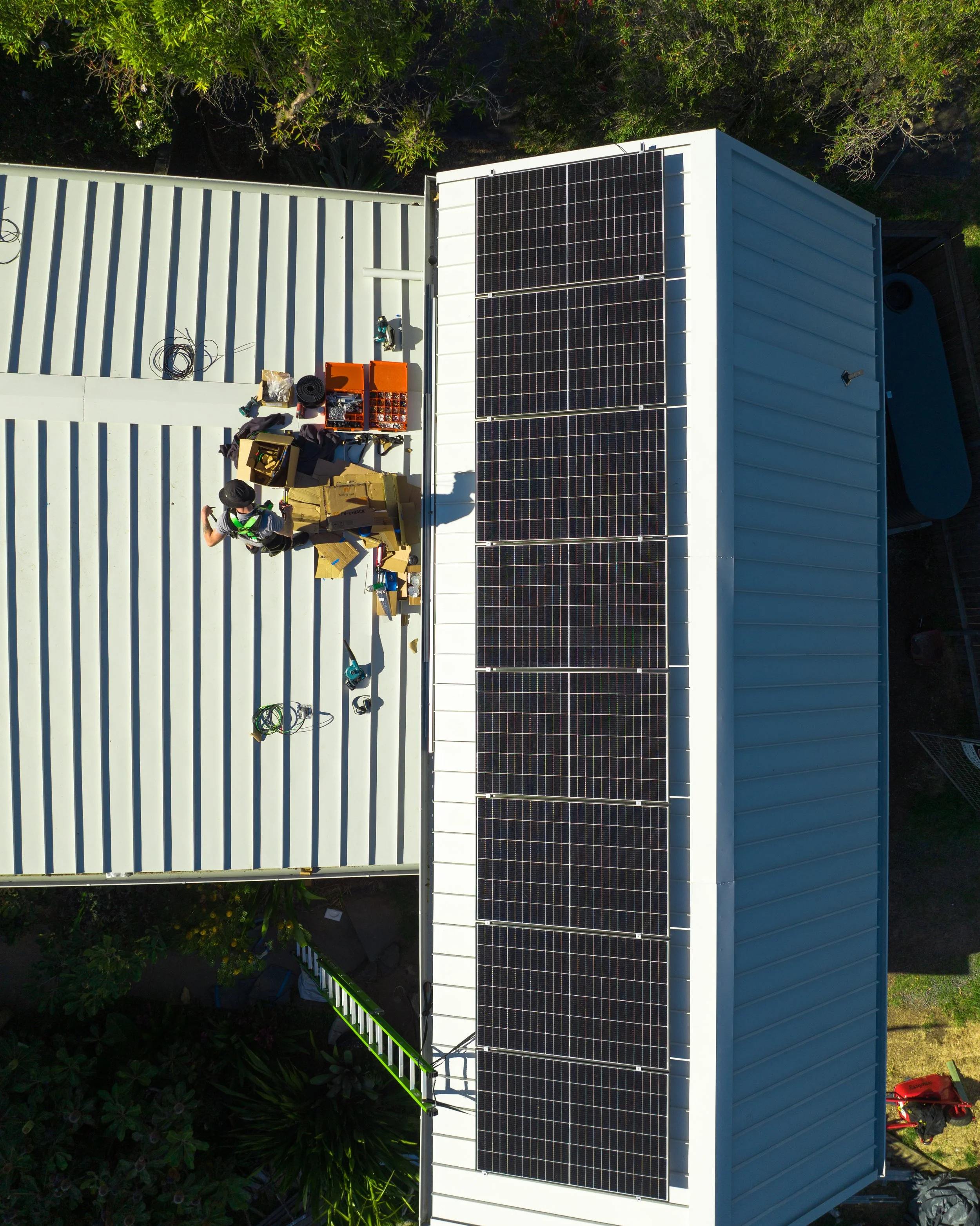 An aerial view of a white building rooftop with solar panels installed. There are tools and equipment on the roof, and a person working near the solar panels.