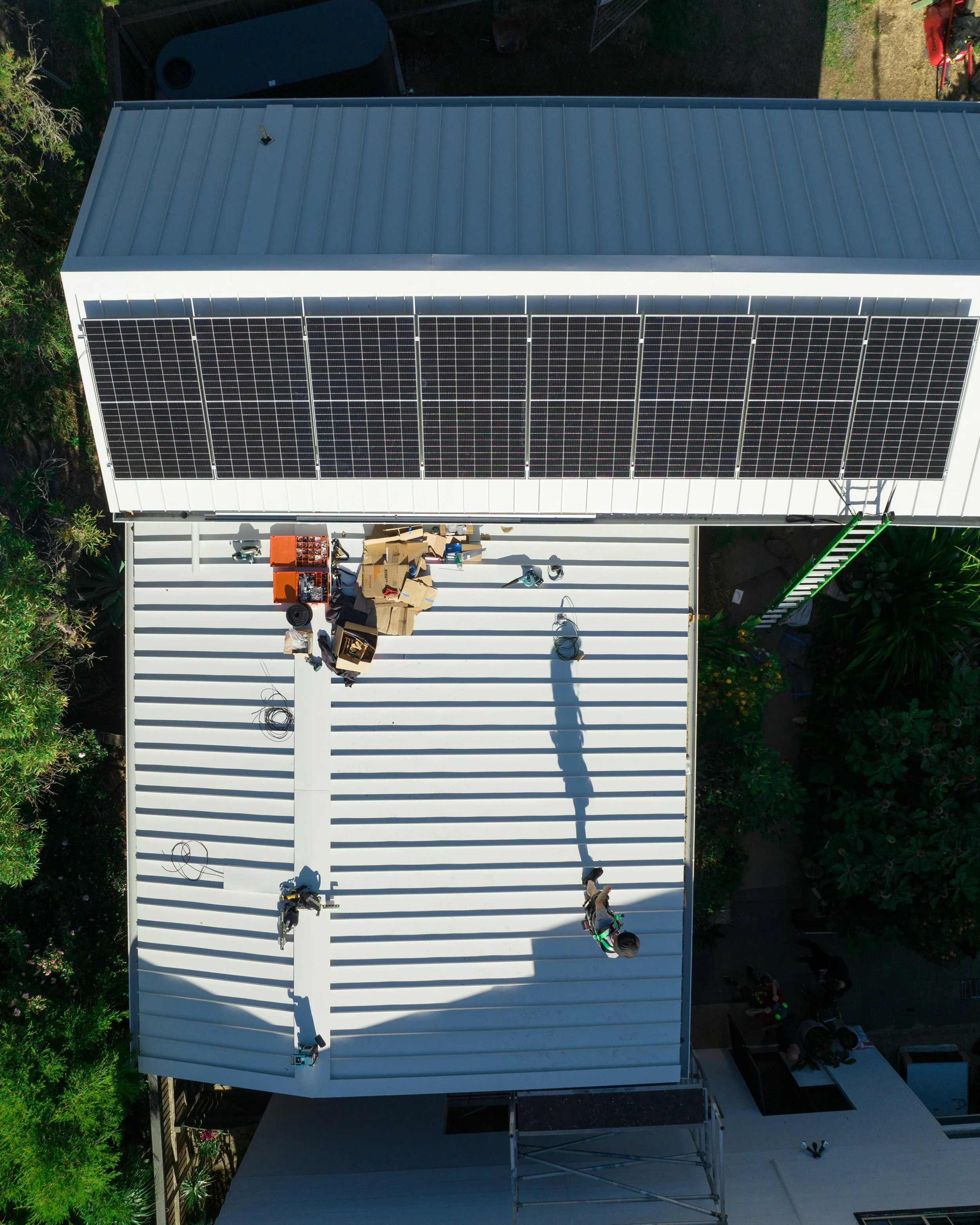 Aerial view of a building's white metal roof with solar panels installed on one side, surrounded by greenery and outdoor equipment.
