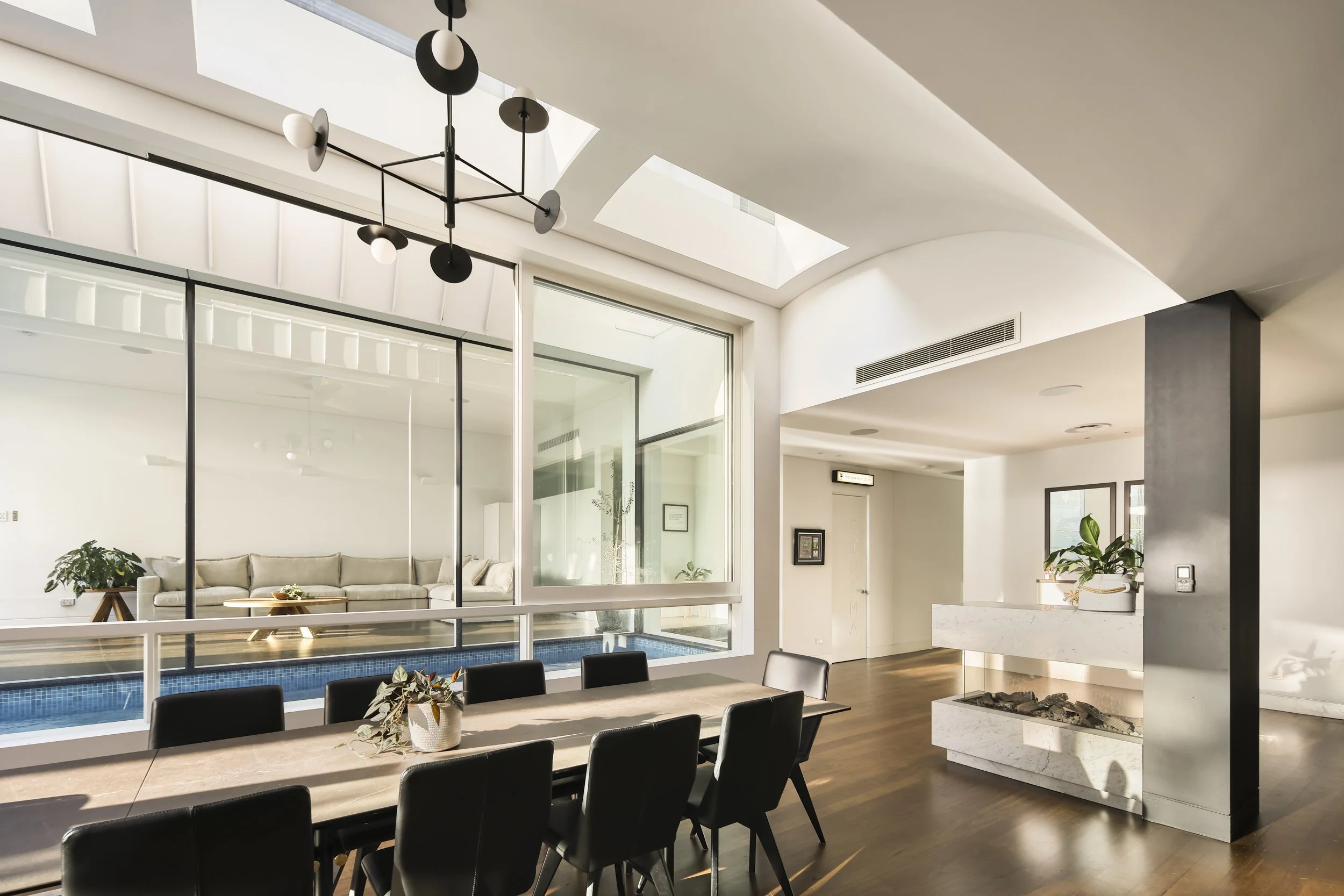 Modern interior with a large dining table surrounded by black chairs, a white marble fireplace with decorative stones, large windows, and skylights letting in natural light, plants, and contemporary lighting fixtures.