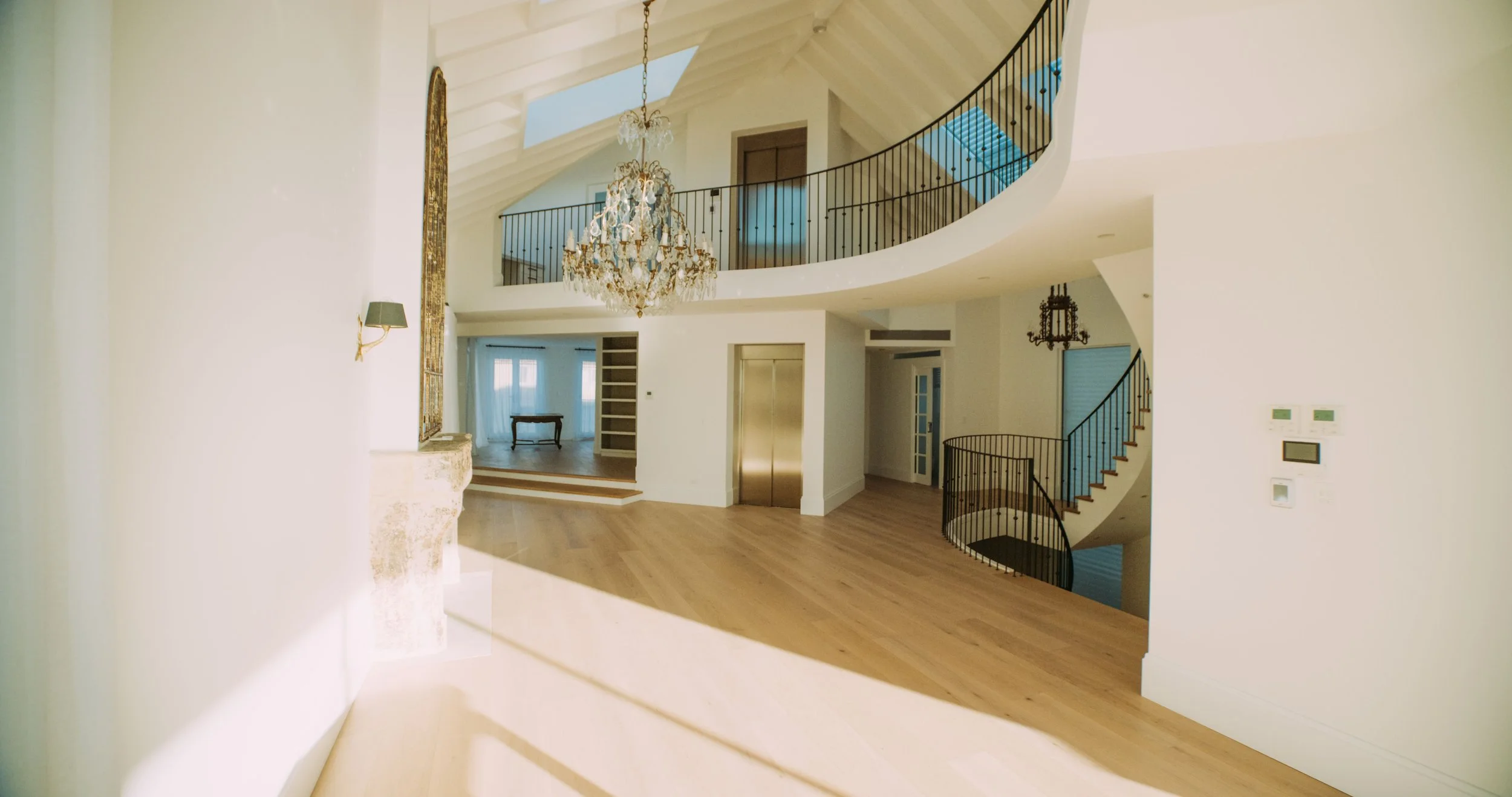 Interior view of a spacious, modern house with high ceilings, wooden floors, a grand chandelier, a staircase with black railings, an elevator, and large windows.