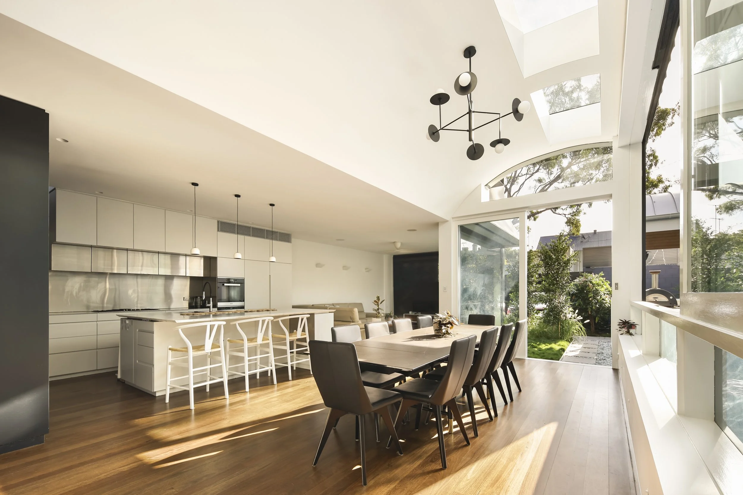 A modern, open-concept kitchen and dining area with a high, curved ceiling and large windows. The space features a kitchen island with white bar stools, a long wooden dining table surrounded by black chairs, and pendant and chandelier lighting. Outsi