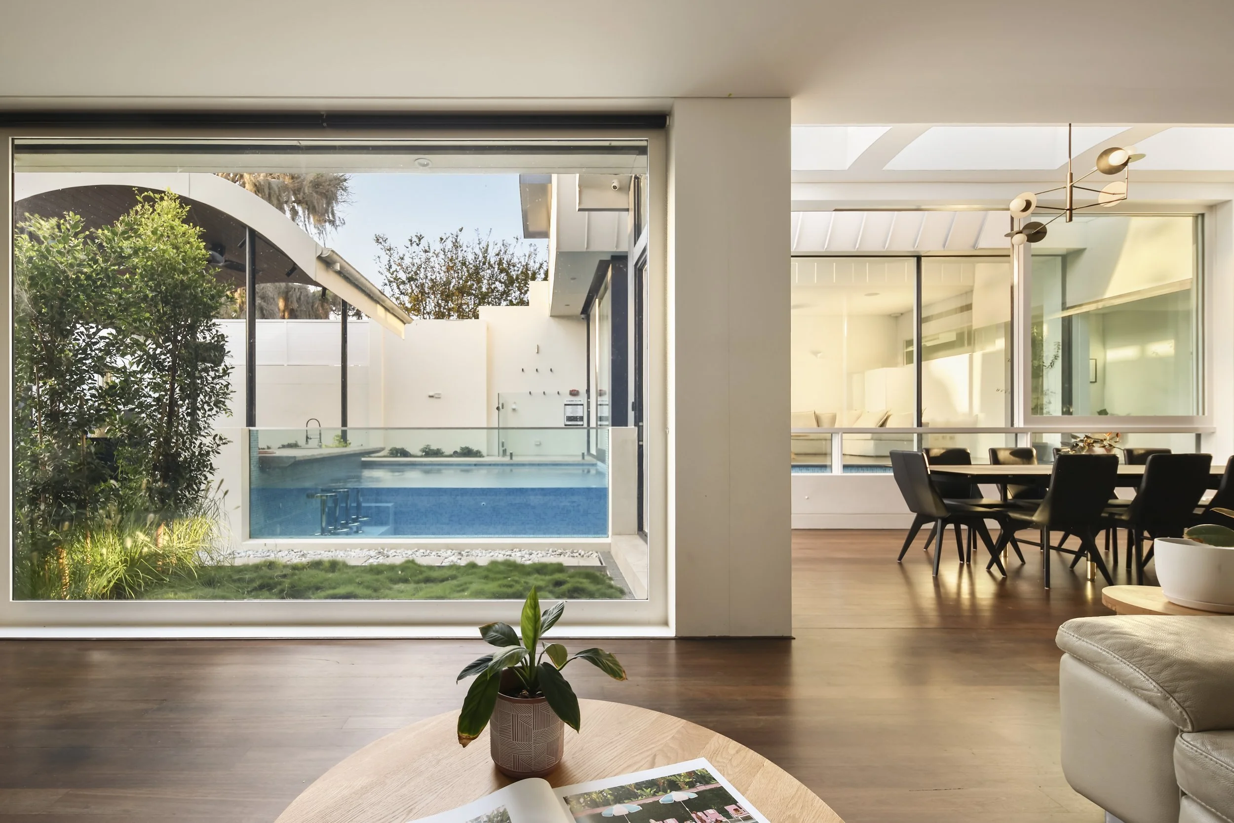 Modern living room with large sliding glass door opening to backyard with pool, outdoor seating, and greenery, interior features a dining area and a potted plant on a wooden coffee table.