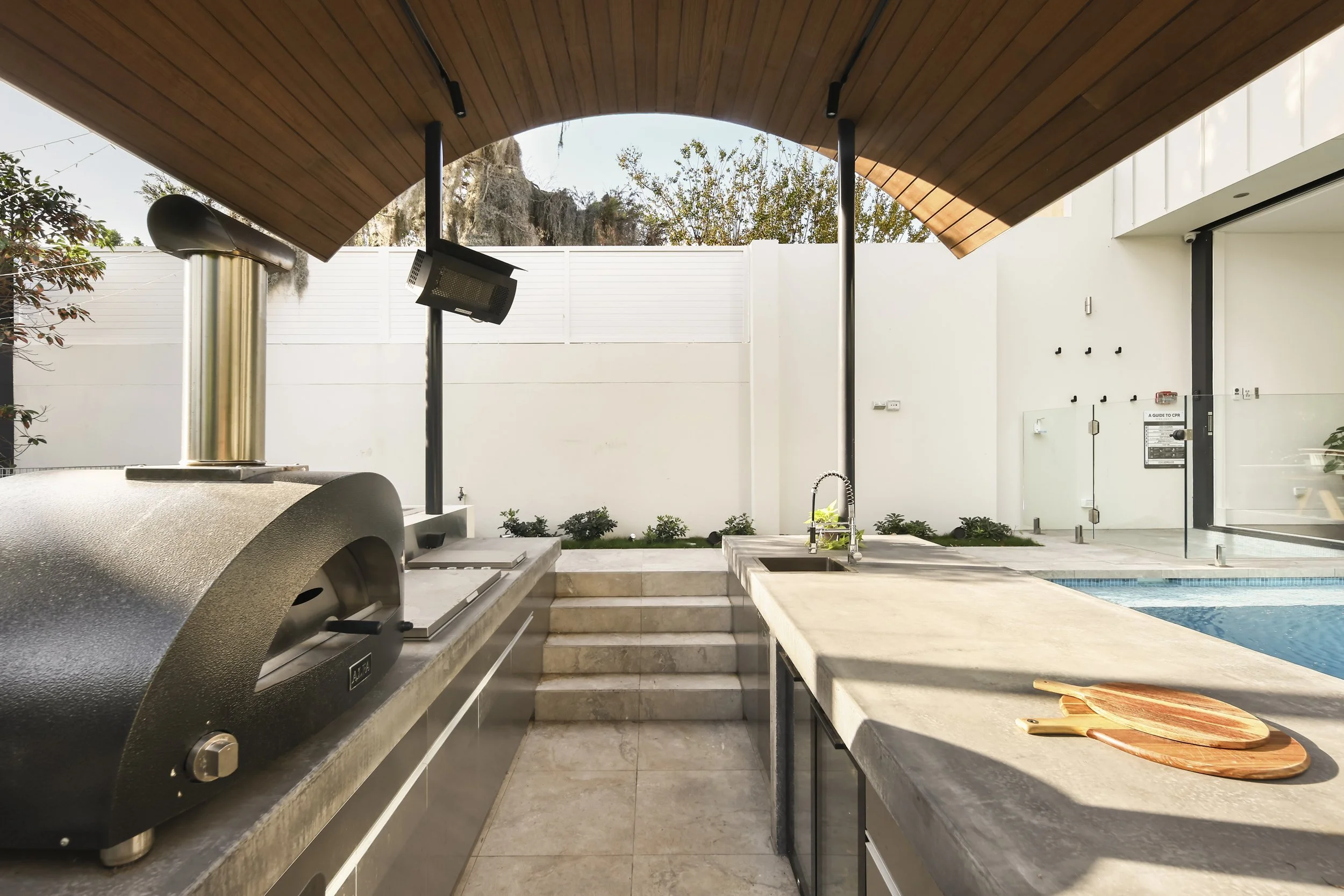 Outdoor kitchen with a barbecue grill, a countertop with wooden cutting boards, a sink, and a swimming pool with steps, surrounded by a white fence and trees, under a curved wooden roof.