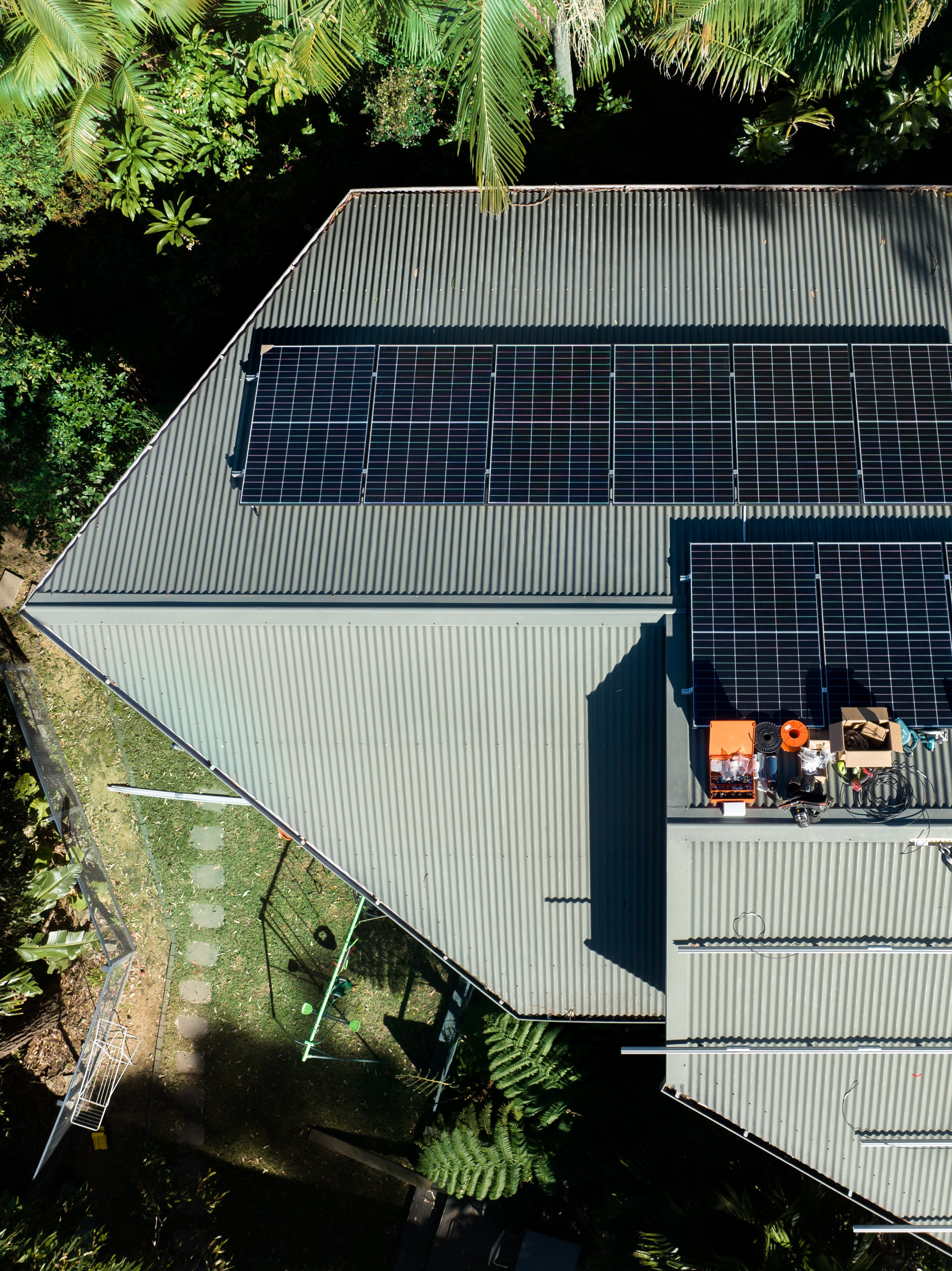 Aerial view of a house with solar panels on the gray metal roof, surrounded by lush green trees and plants.