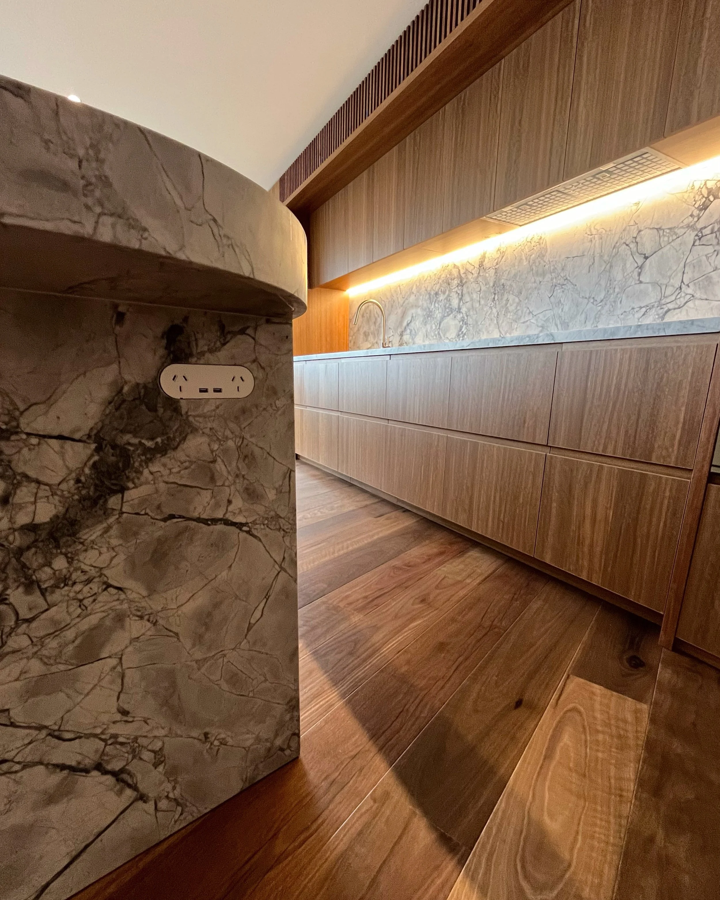 Close-up view of a marble kitchen island with an electrical outlet, beside wooden cabinets and a marble backsplash with ambient lighting.