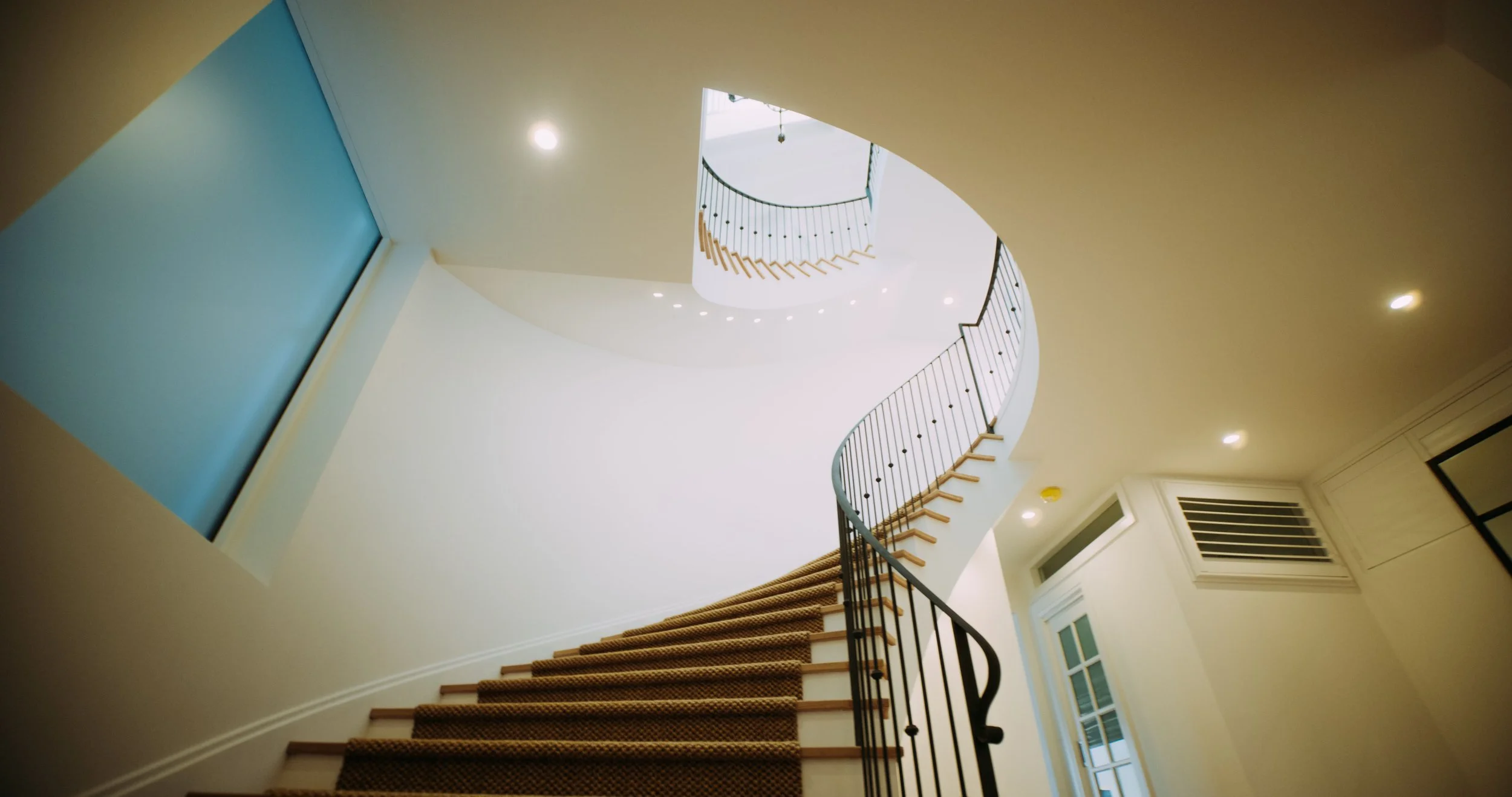 Interior view of a stairwell with a spiral staircase visible from below, featuring wooden steps, black metal railings, and white walls with recessed lighting.