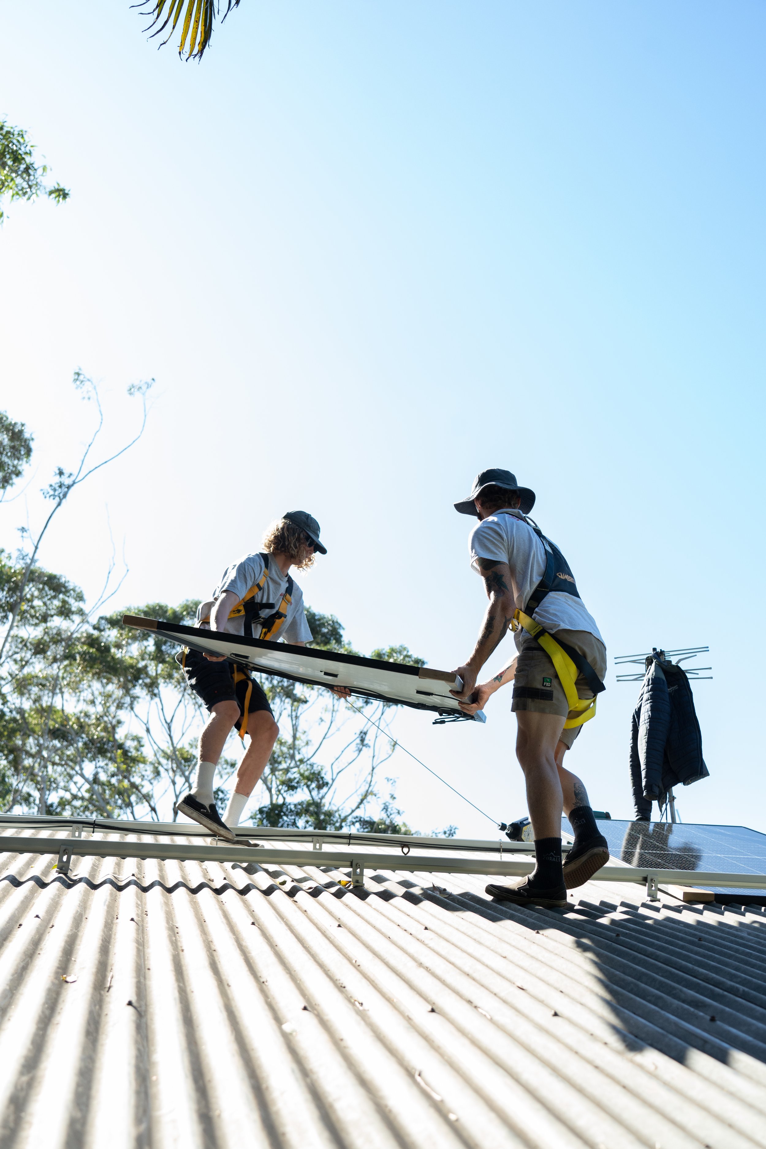 Two people on a roof installing solar panels, with a clear sky and trees in the background.