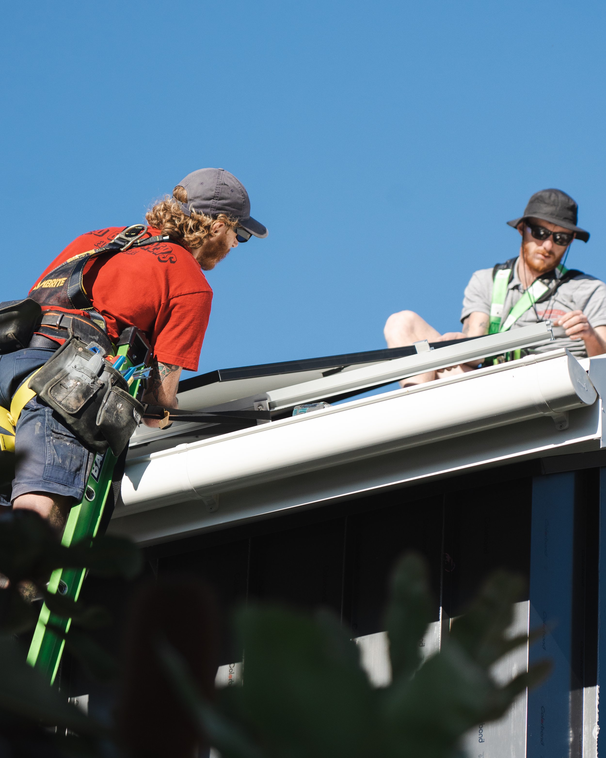 Two men wearing hats and sunglasses working on a roof under a clear blue sky, surrounded by trees.