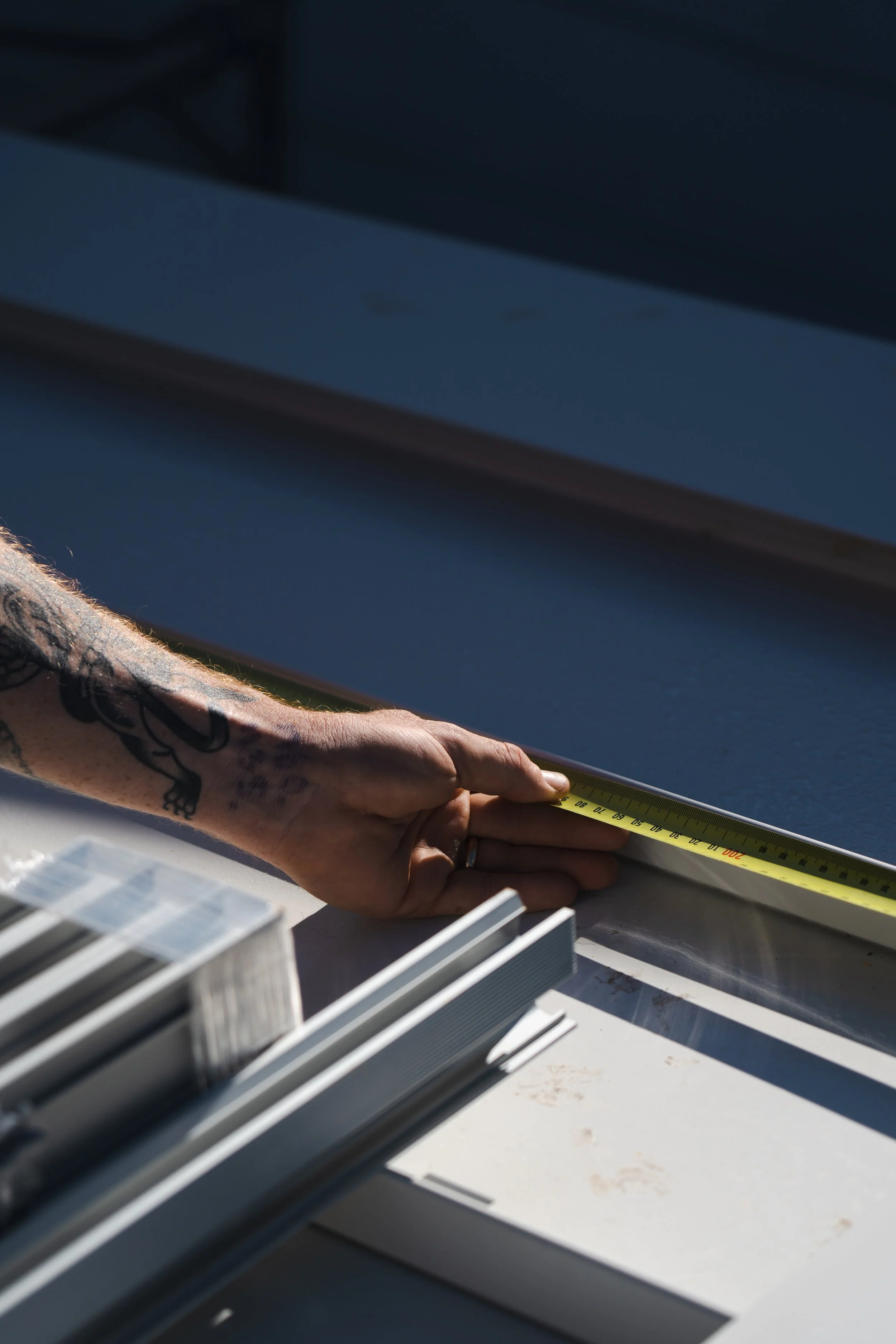A person's hand with tattoos holding a measuring tape, measuring a white window frame, with white window sashes in the foreground.