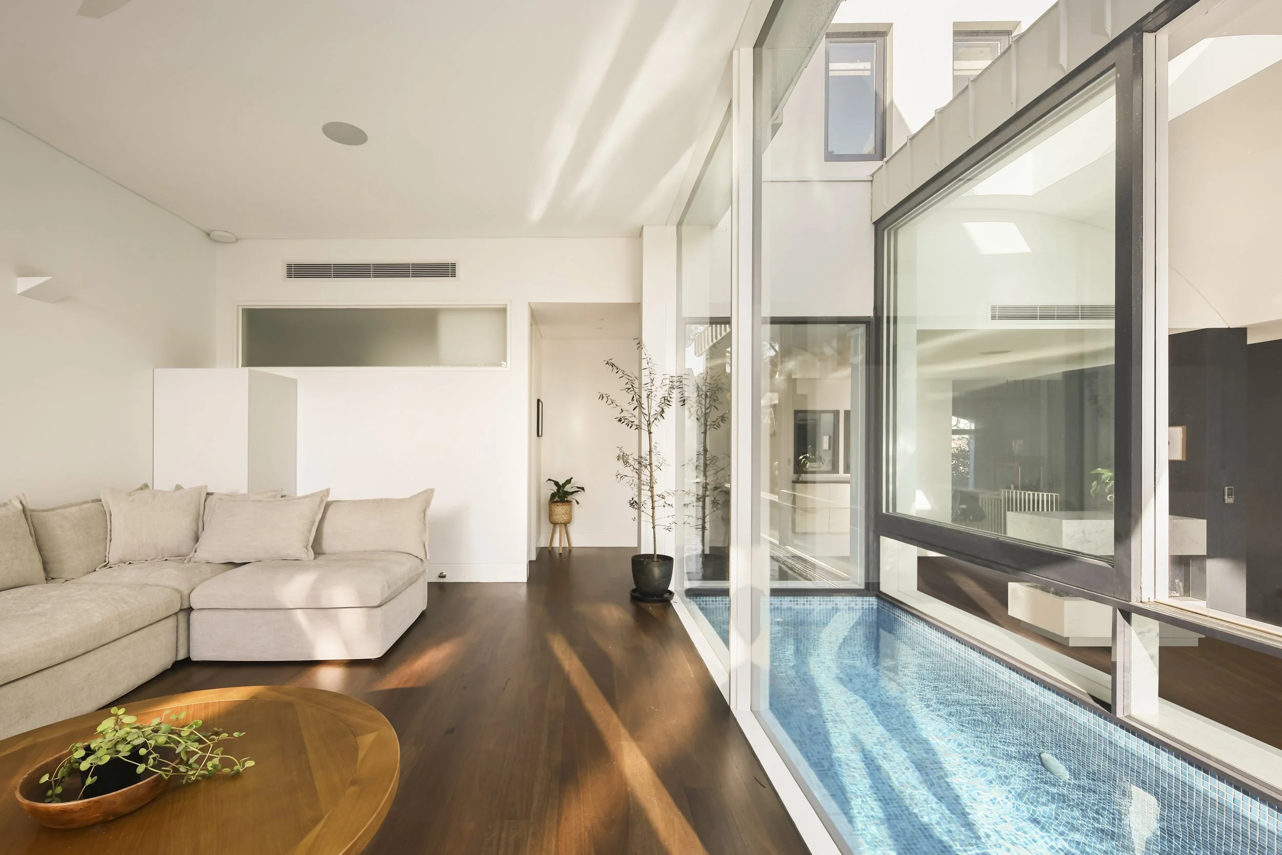 Modern living room with beige sectional sofa, dark hardwood floors, large windows letting in natural light, and a small indoor pool with a glass wall.