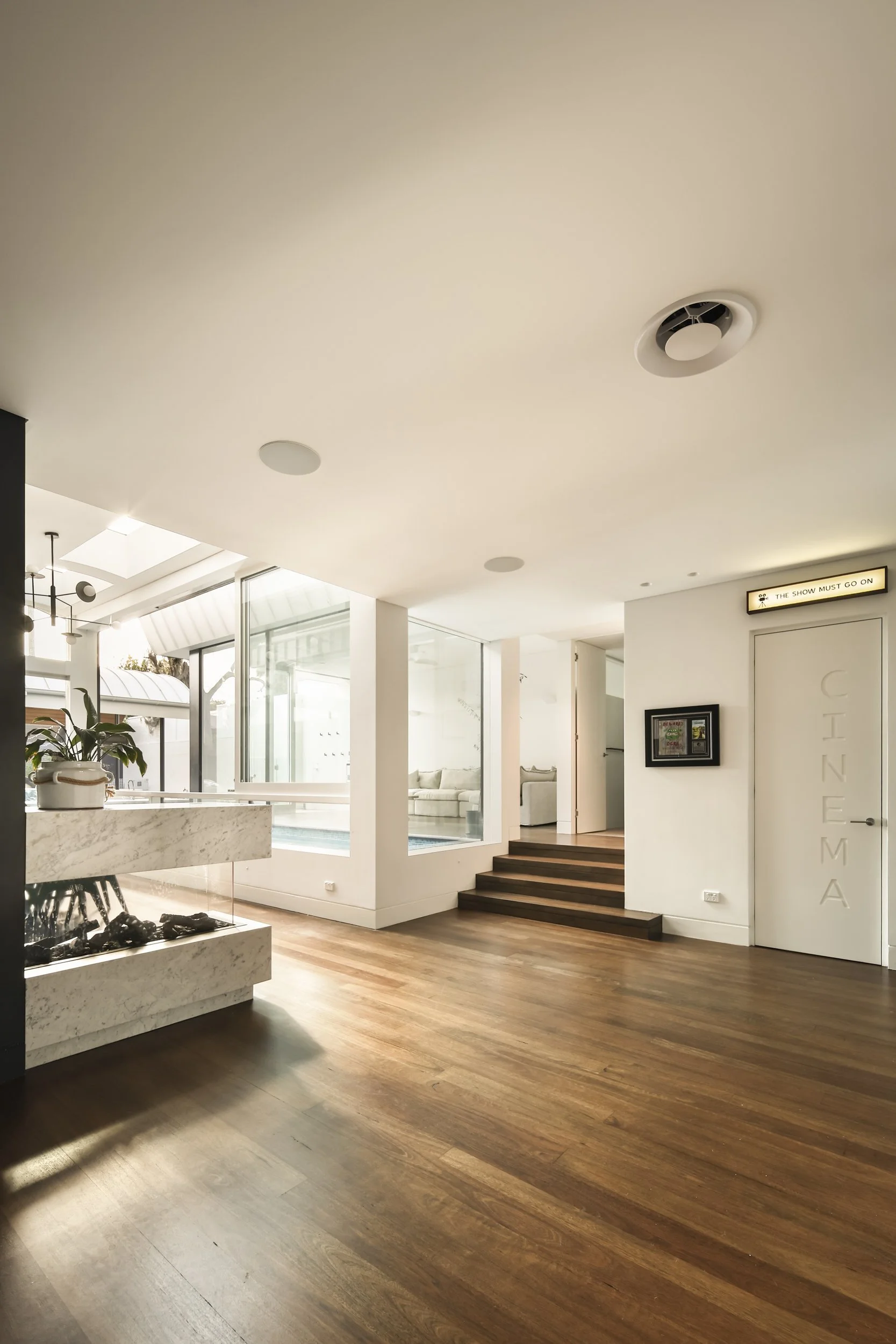A modern interior space with wooden flooring, stairs, a white ceiling with round recessed lights and a ceiling vent, large windows, a marble fireplace, and white walls with framed pictures and a sign reading 'The Show Must Go On'.