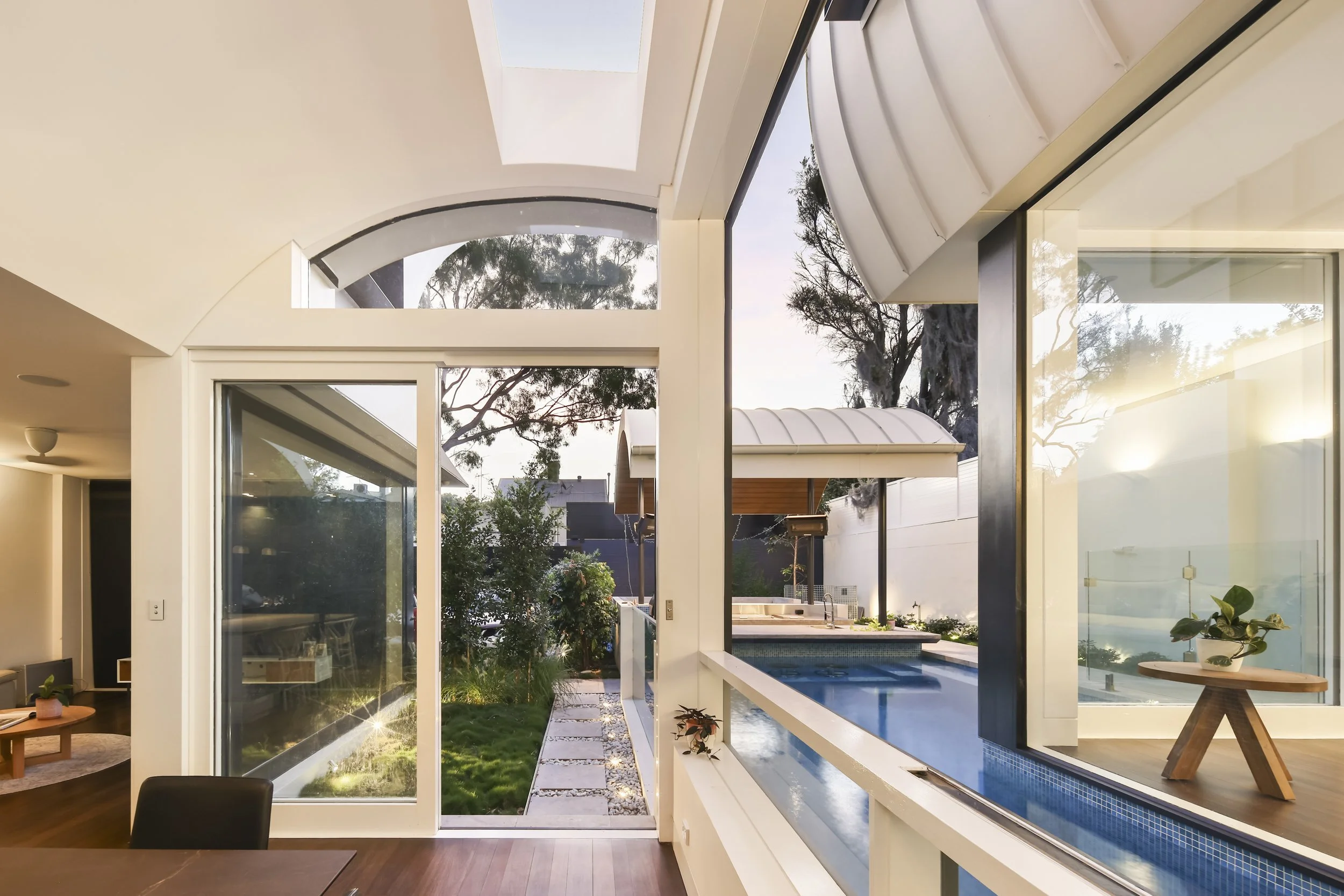 Interior view of a modern house showing large floor-to-ceiling glass windows and doors, with a backyard featuring a small pool, patio, and outdoor seating area, during daylight.