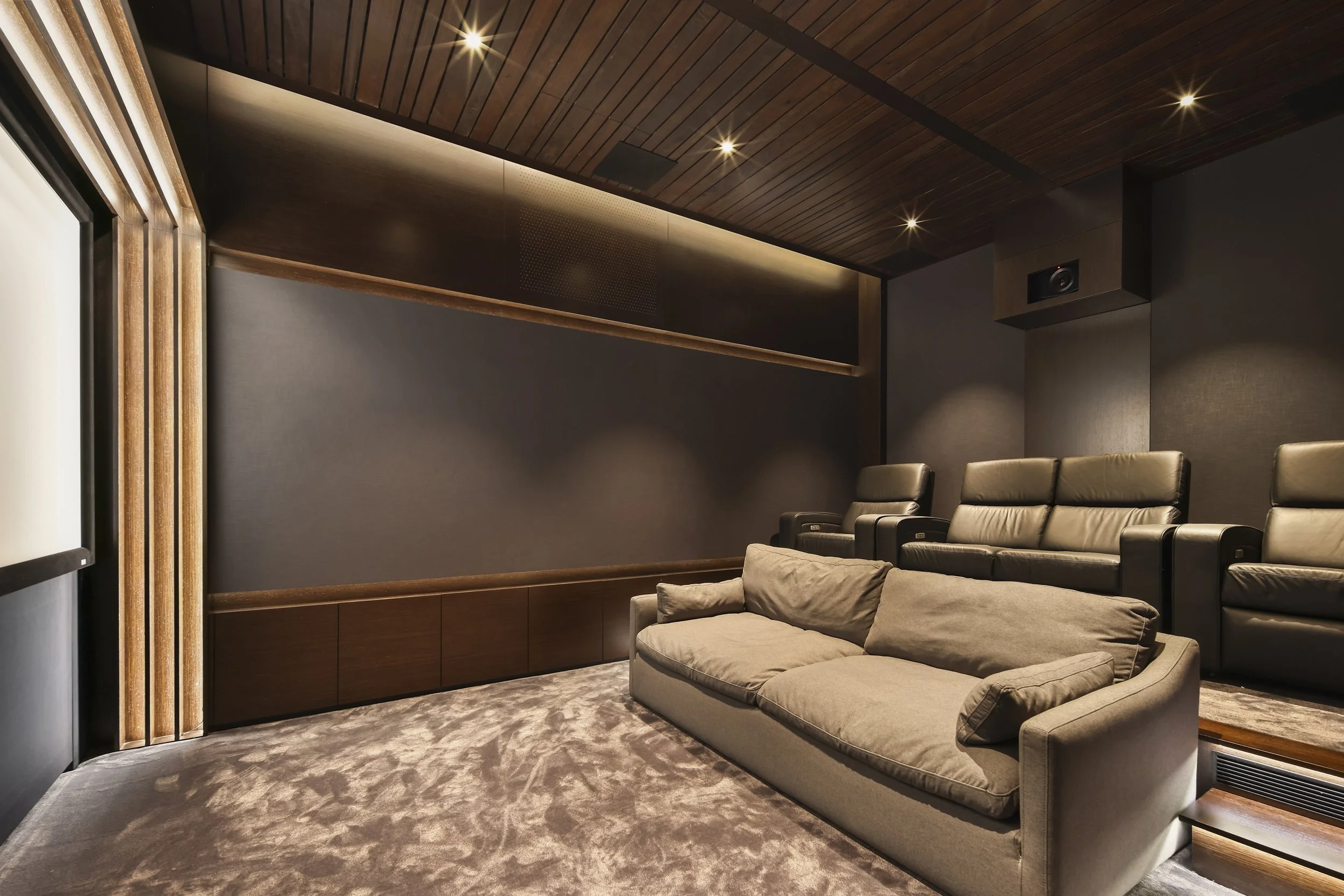A cozy home theater room with a beige sofa and reclining chairs. The ceiling and wall are wood-paneled, with soft recessed lighting and a dark wall with soundproofing material. There is a flat screen television mounted on the wall.