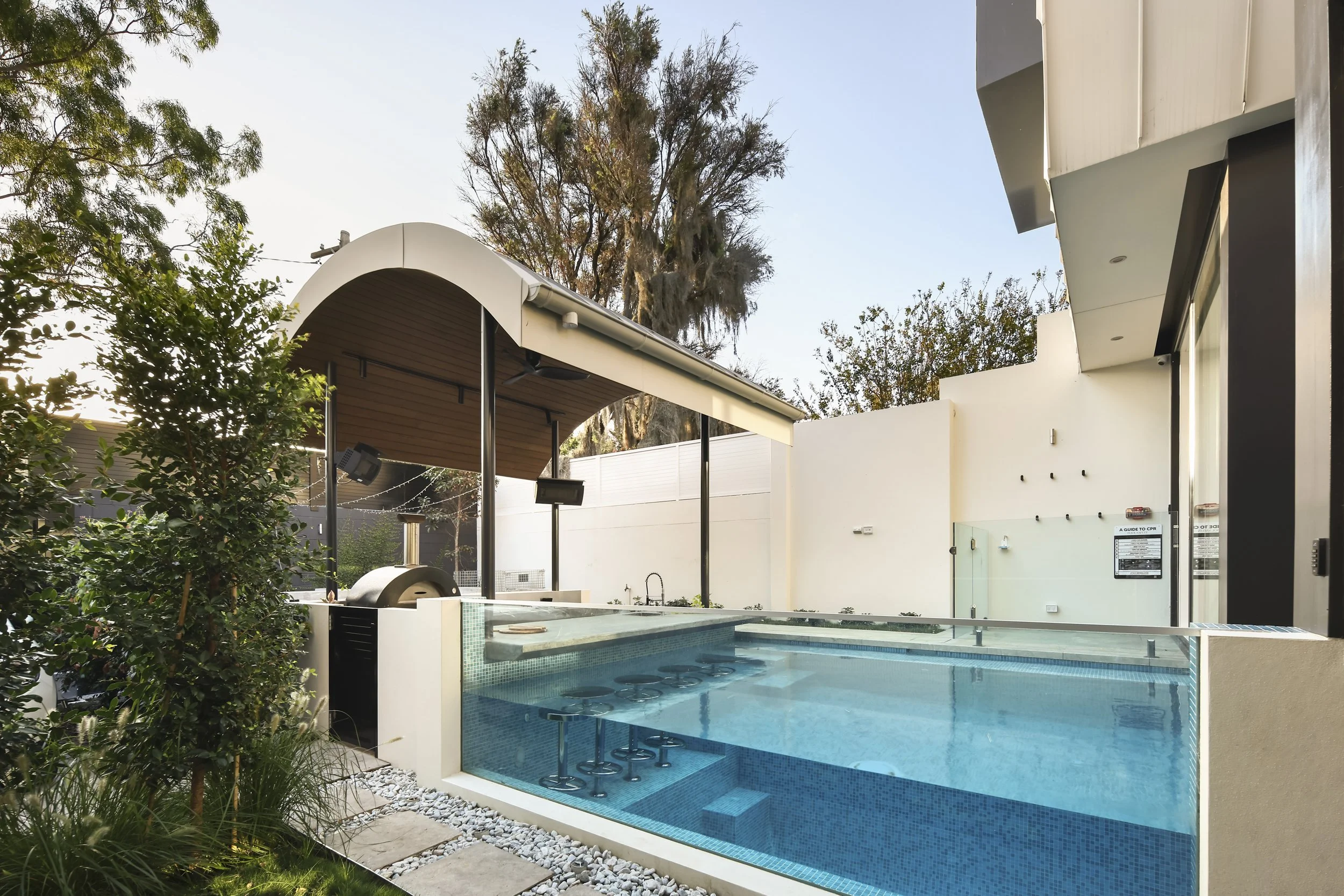 Modern backyard with a swimming pool, barstools, and a covered patio area. Green bushes and trees surround the yard, with tall trees in the background and a clear sky above.