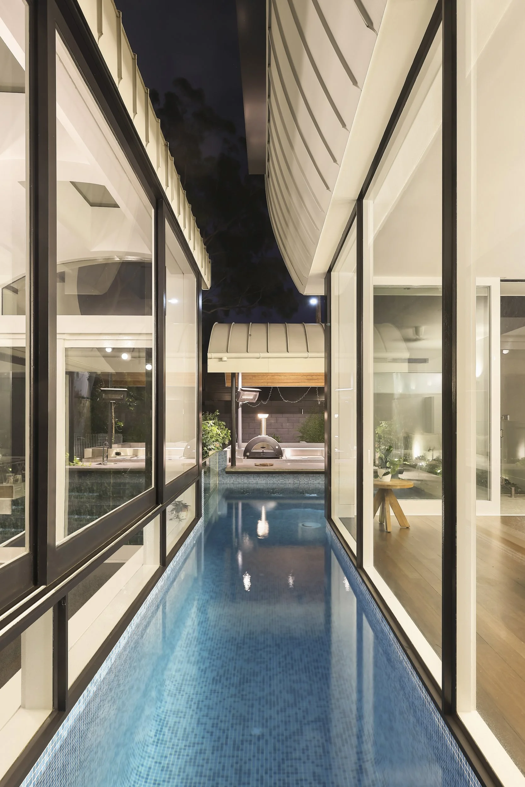View of a modern indoor swimming pool through glass sliding doors at night. The pool has blue mosaic tiles and warm lighting, with a hot tub or small pool at the end, and potted plants and furniture visible inside.