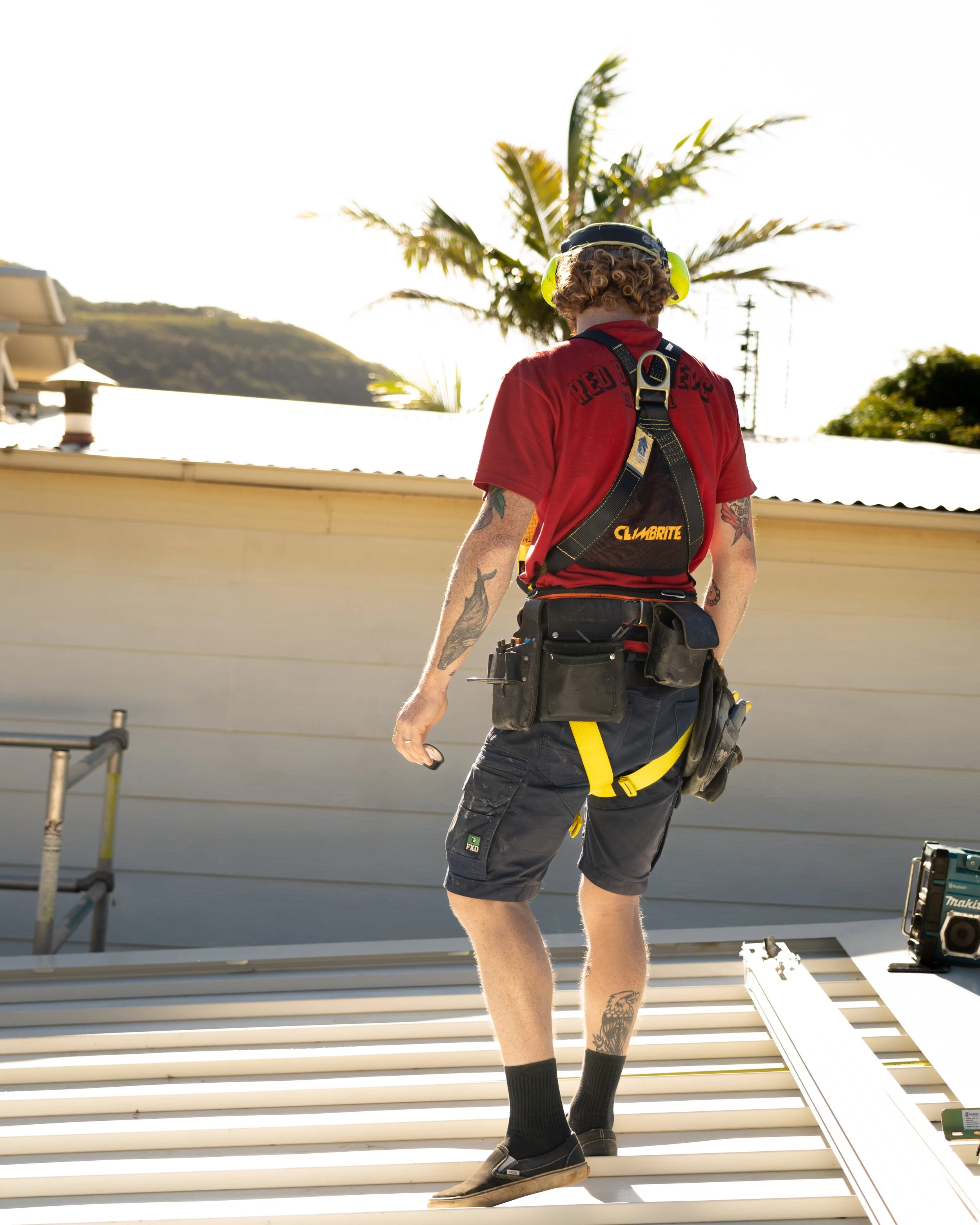 A person with curly hair and tattoos on their arm, dressed in a red shirt, black shorts, and black shoes, is wearing a safety harness and yellow ear protection, and is working on a construction or maintenance project outdoors near a roof with a palm 