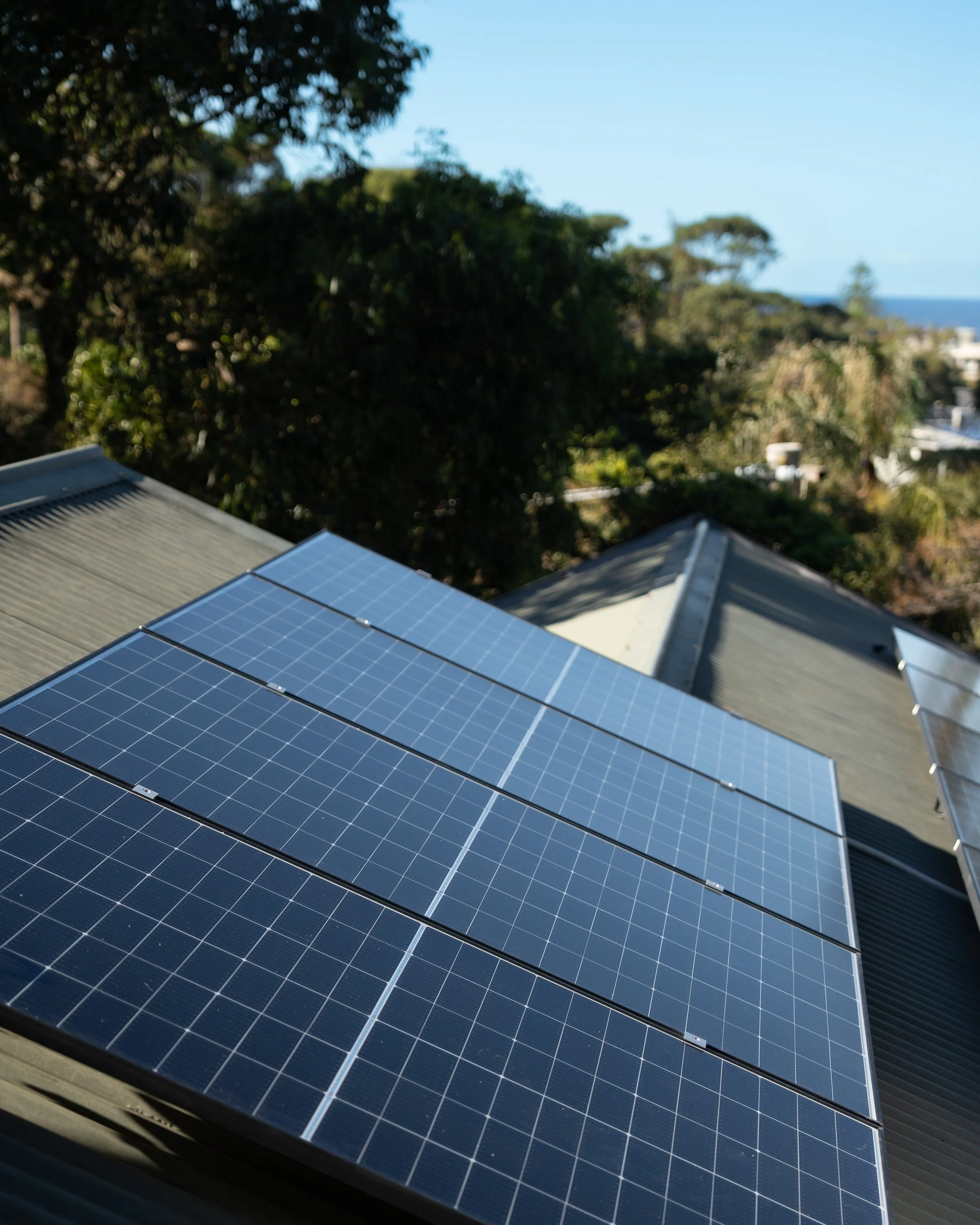 Solar panels installed on a metallic roof with a background of green trees and a clear blue sky.