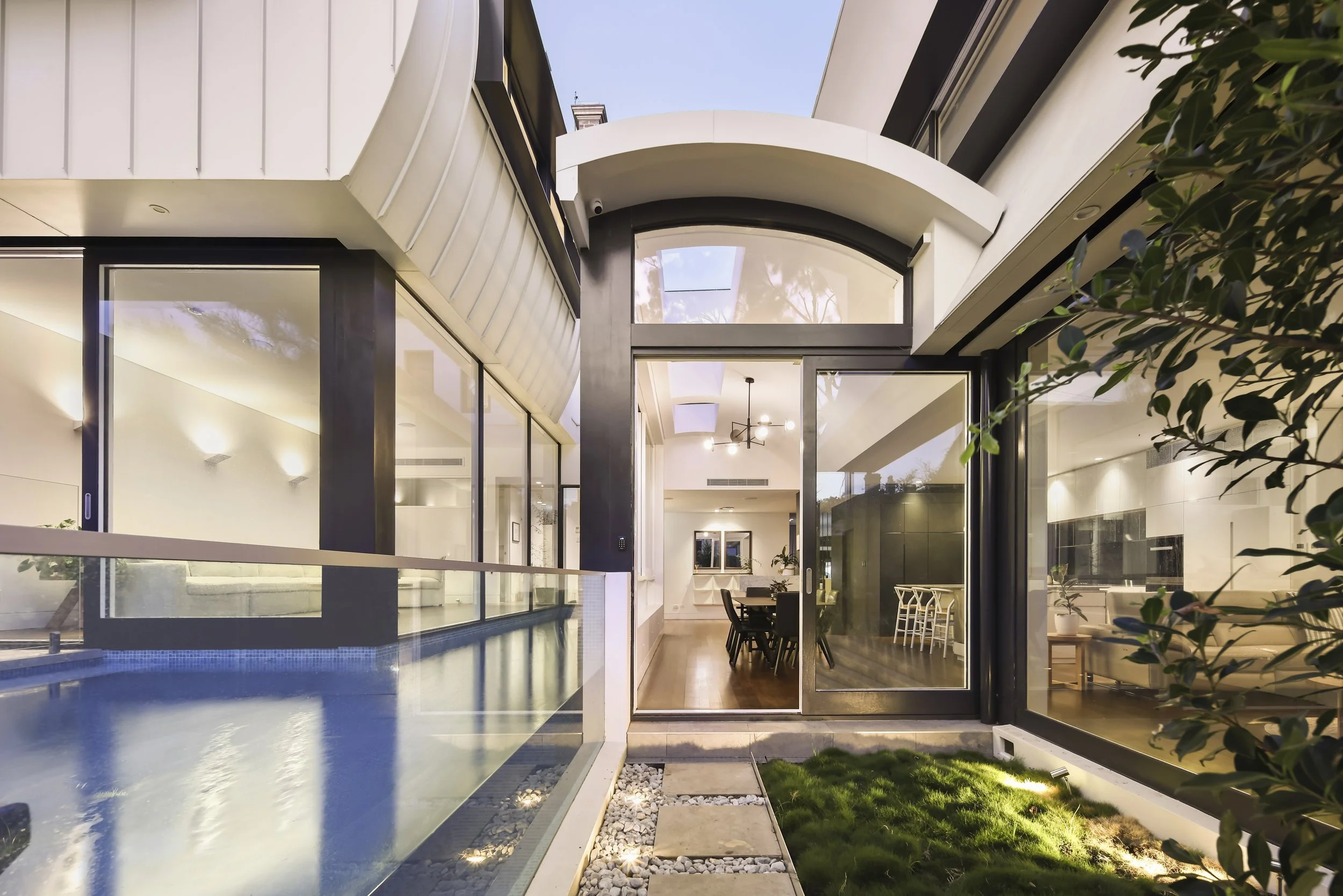 Modern house with a backyard pool, large glass windows, and sliding doors. The interior features a dining area and living space with contemporary furniture and lighting. Exterior includes a small lawn and decorative rocks.