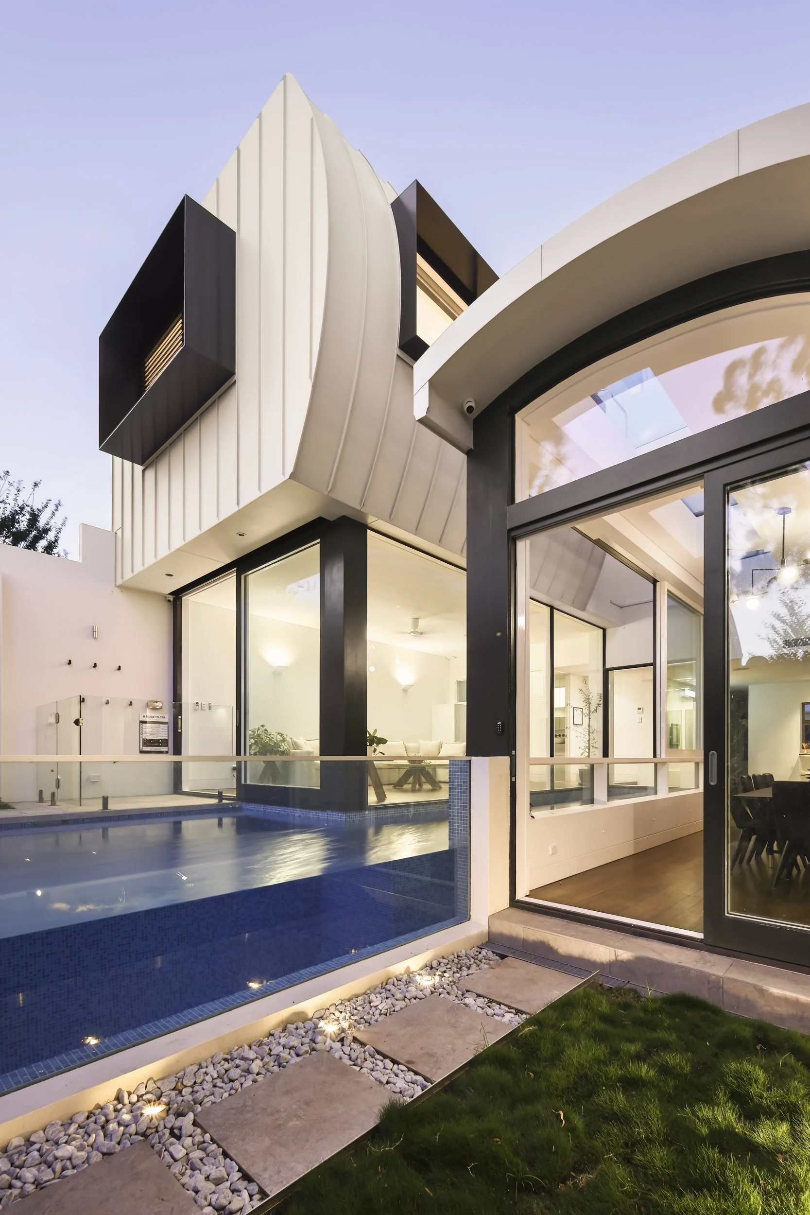 Modern house exterior at dusk with large glass windows, pool, and landscaped yard with rocks and grass.