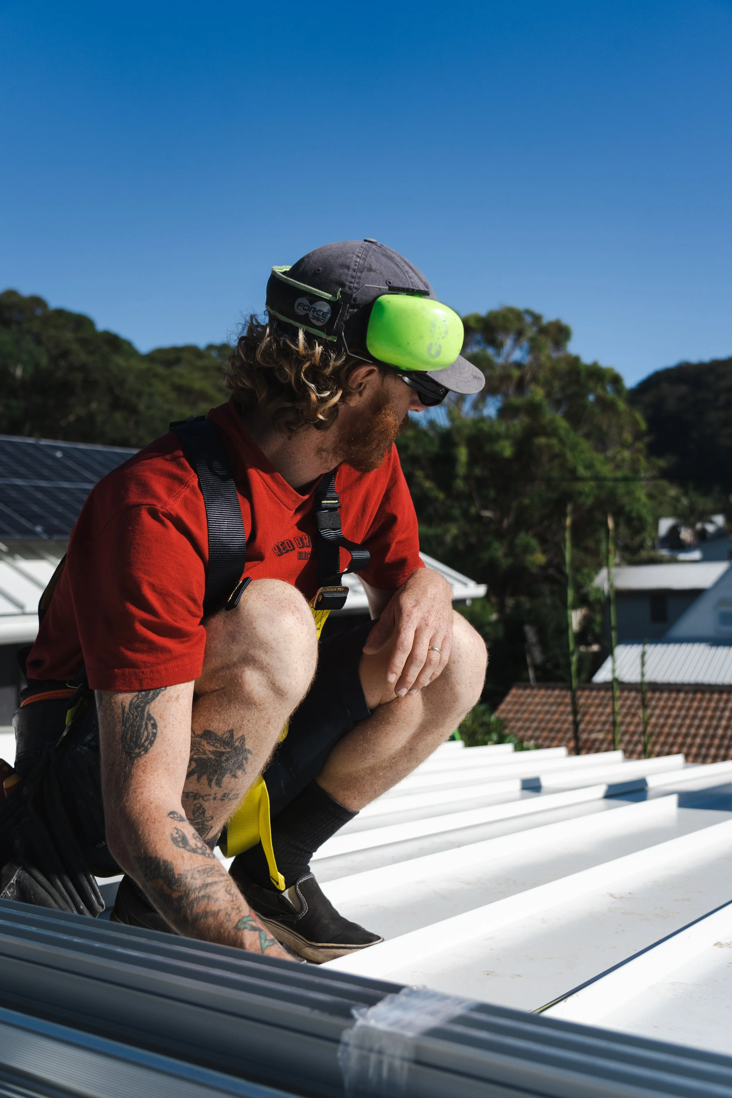 A man with tattoos and a beard, wearing a red shirt, a gray cap, and sunglasses, is crouched on a white rooftop during daytime, wearing green hearing protection and a yellow safety harness, with a background of trees, houses, and a clear blue sky.