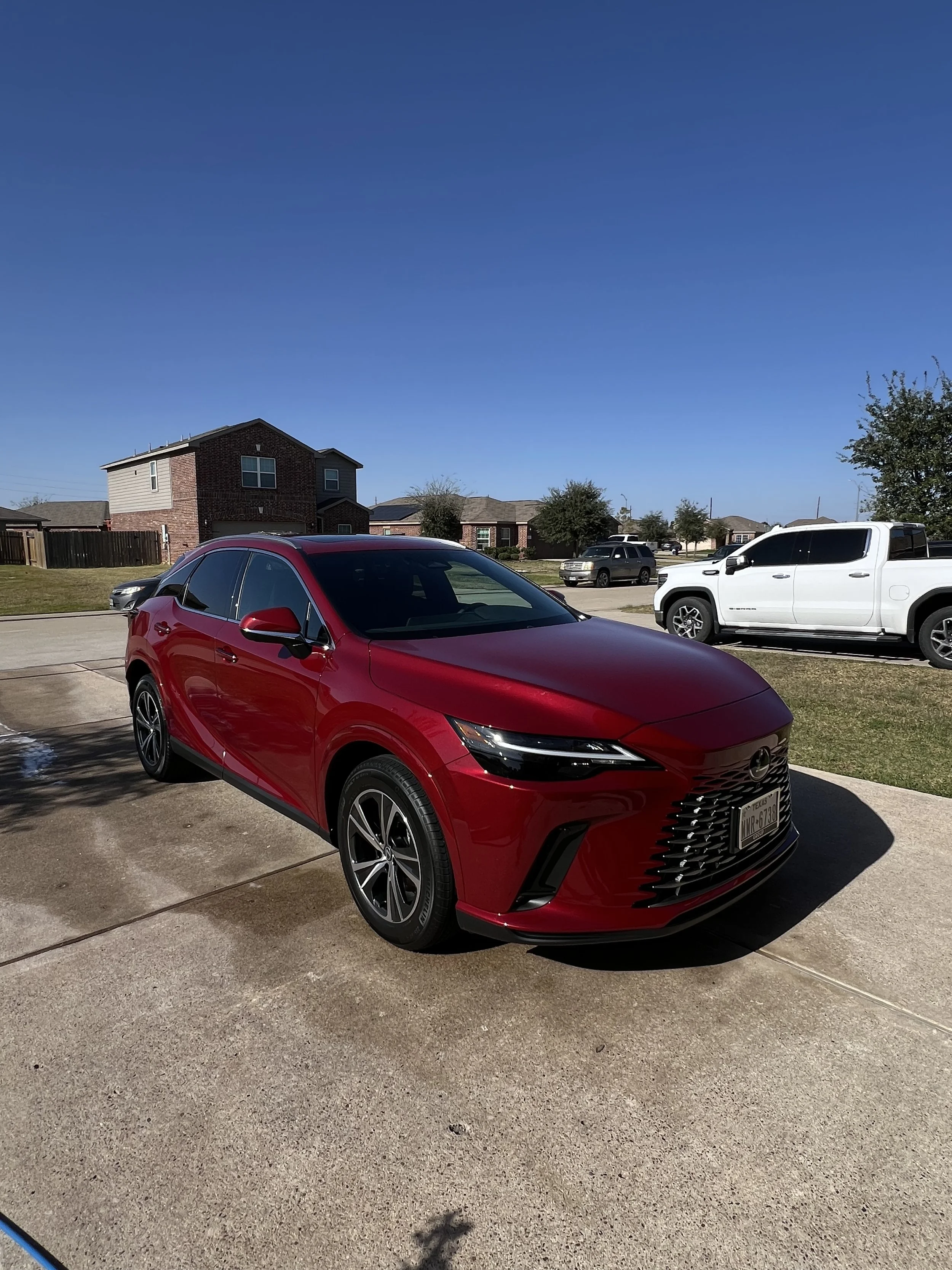 A red Mazda crossover SUV parked on a driveway in front of a suburban house, with other cars and houses visible in the background under a clear blue sky.