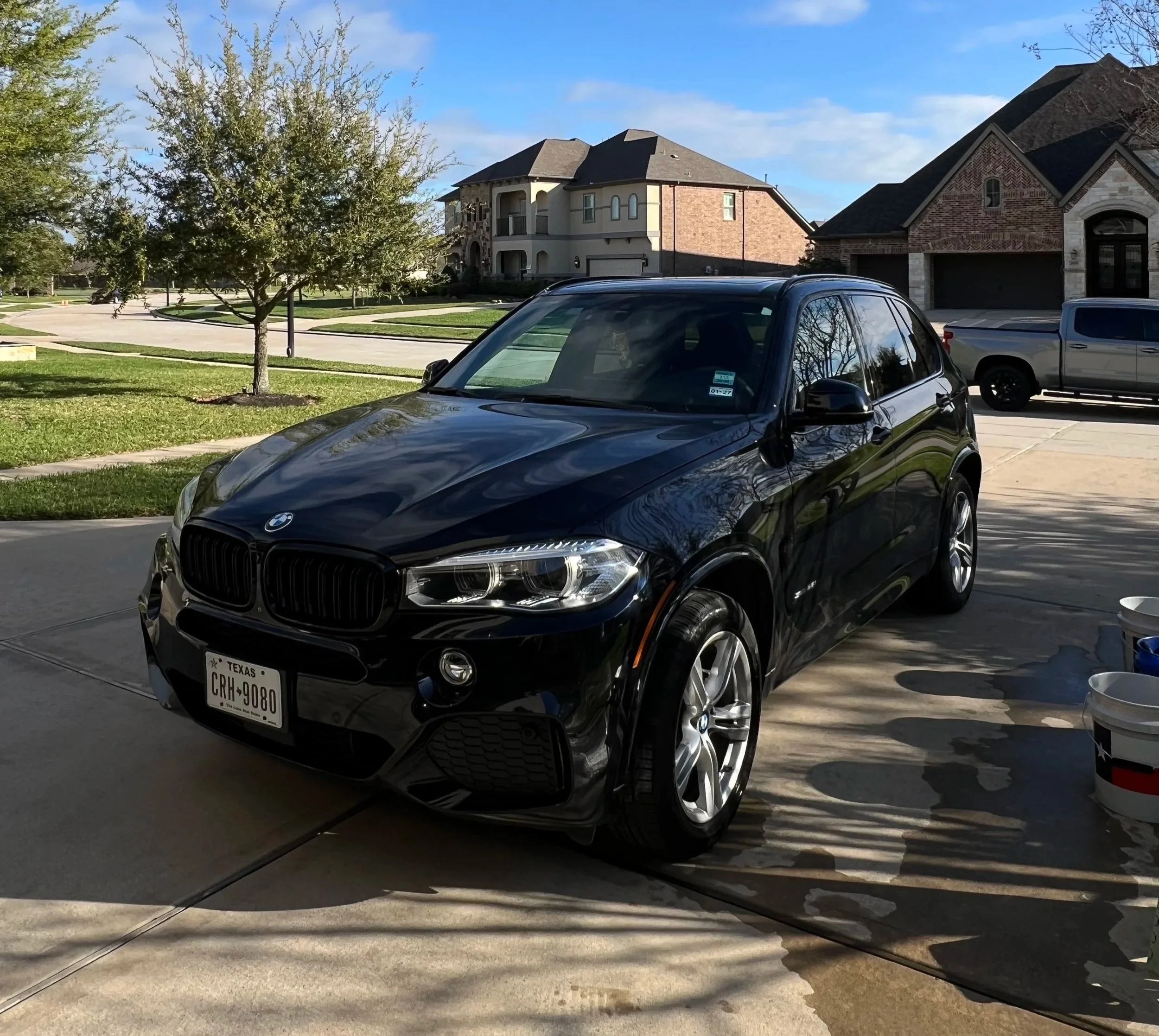 A suburban neighborhood street scene showing a black BMW SUV parked in a driveway, other cars parked along the street, and houses with brick and stone facades. There are trees with green leaves and a clear blue sky.