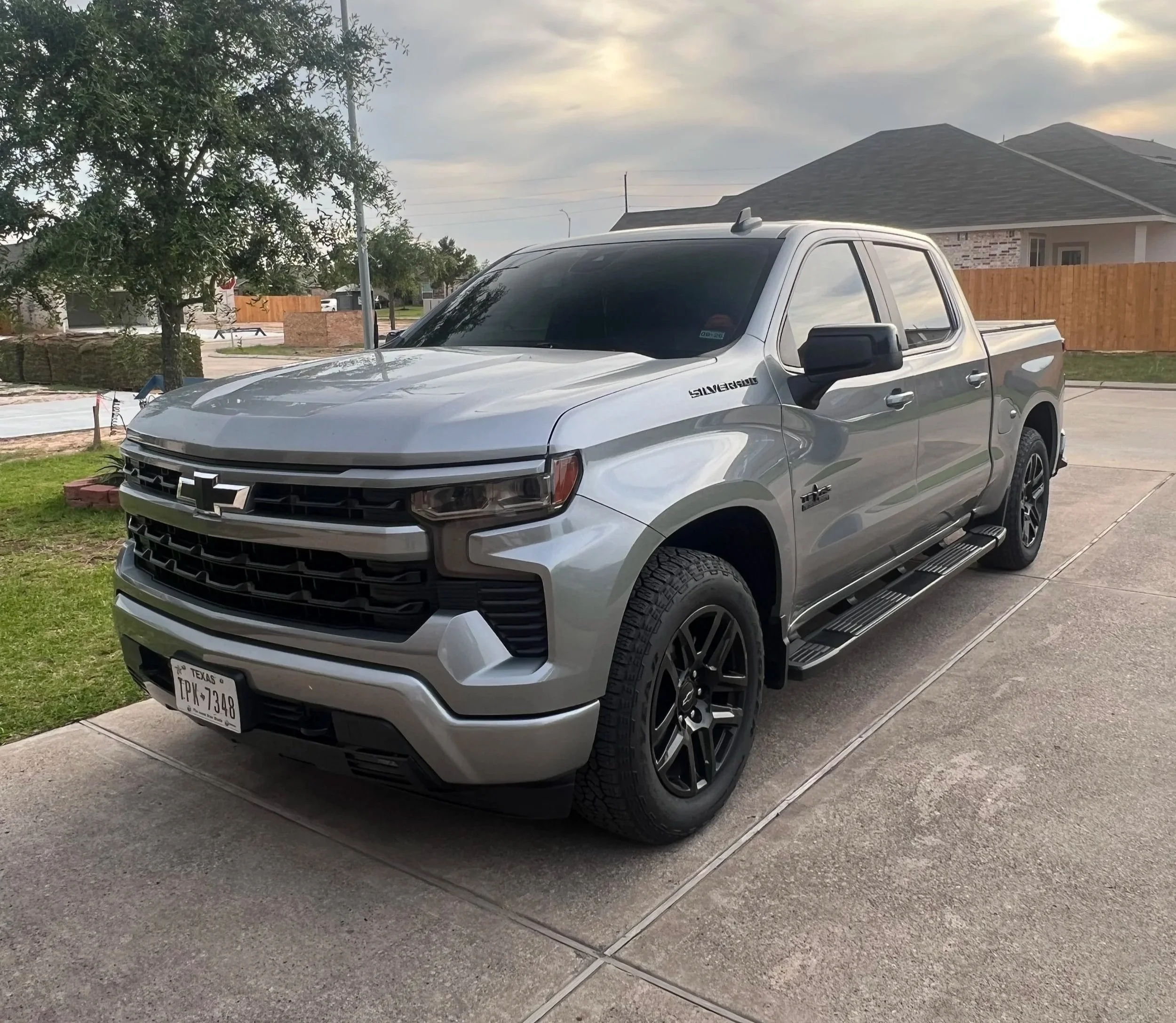 A silver Chevrolet Silverado pickup truck parked on a residential driveway with a house, wooden fence, trees, and cloudy sky in the background.