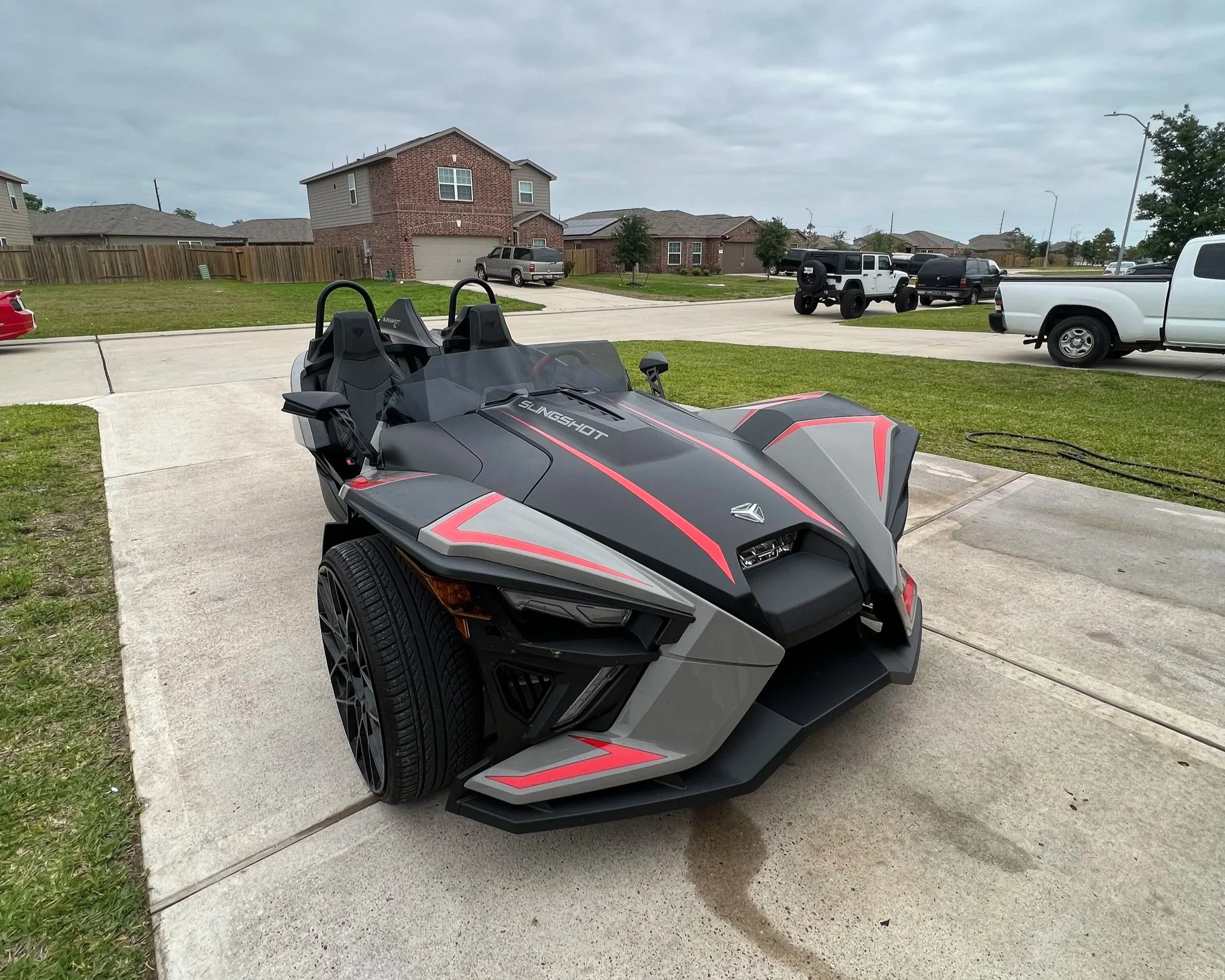 A parking lot in a suburban neighborhood with multiple parked cars including a black and red Polaris Slingshot in the foreground, several SUVs and pickup trucks, and houses with brick and siding exteriors in the background.